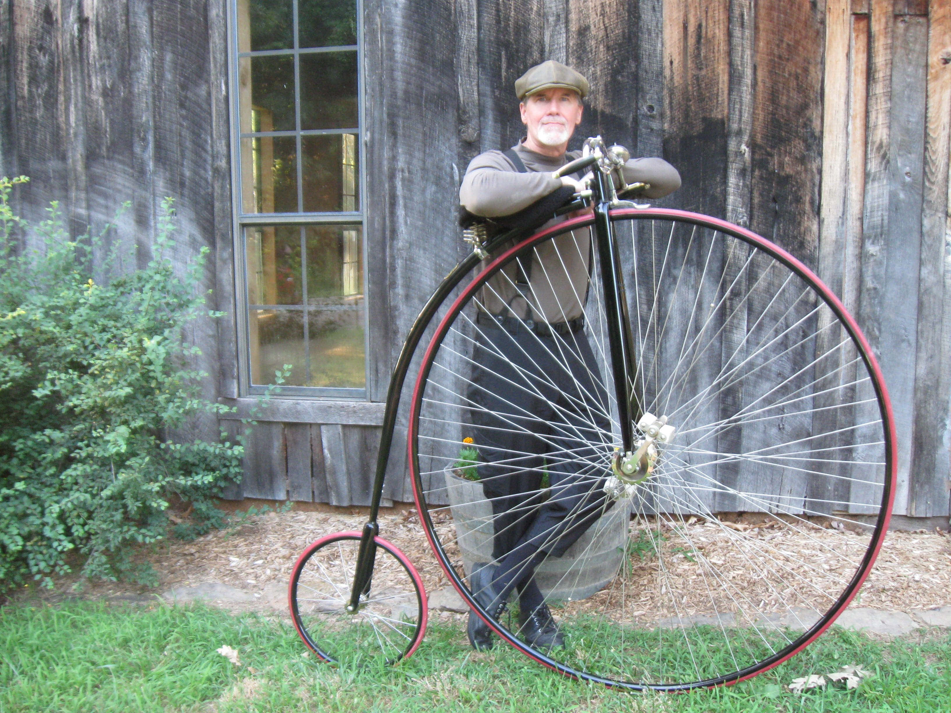 Bicycle 桌布 壁紙 — 一名身穿復古服裝的男子，自豪地站在一輛經典高輪單車旁，背景是風化的木造建築。