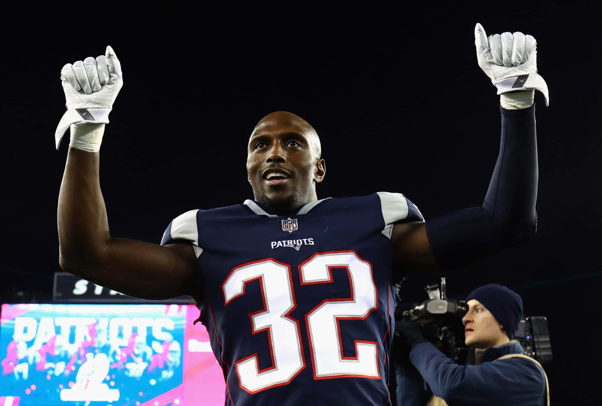 Devin Mccourty 桌布 壁紙 — 一名身穿愛國者隊32號球衣的美式足球員在球場燈光下高舉戴著手套的雙拳慶祝勝利。