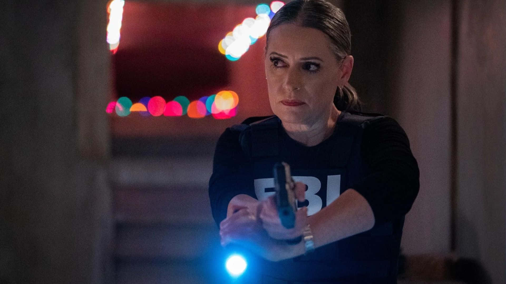 paget brewster 桌布 壁紙 — 一名身穿戰術背心的FBI探員，在昏暗的走廊中瞄準發光的手電筒和手槍，背景有色彩繽紛的散景燈光。