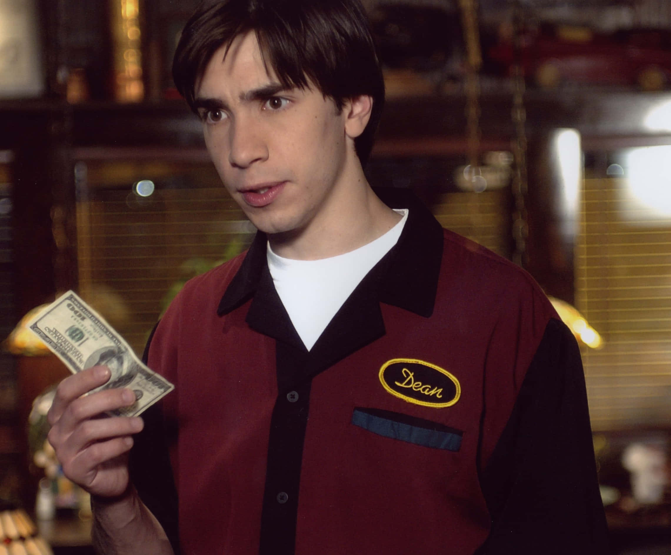 Justin Long 桌布 壁紙 — 一名身穿栗色和黑色制服、名叫Dean的年輕男子，在溫暖燈光的餐廳裡，拿著一張一百美元鈔票，表情驚訝。