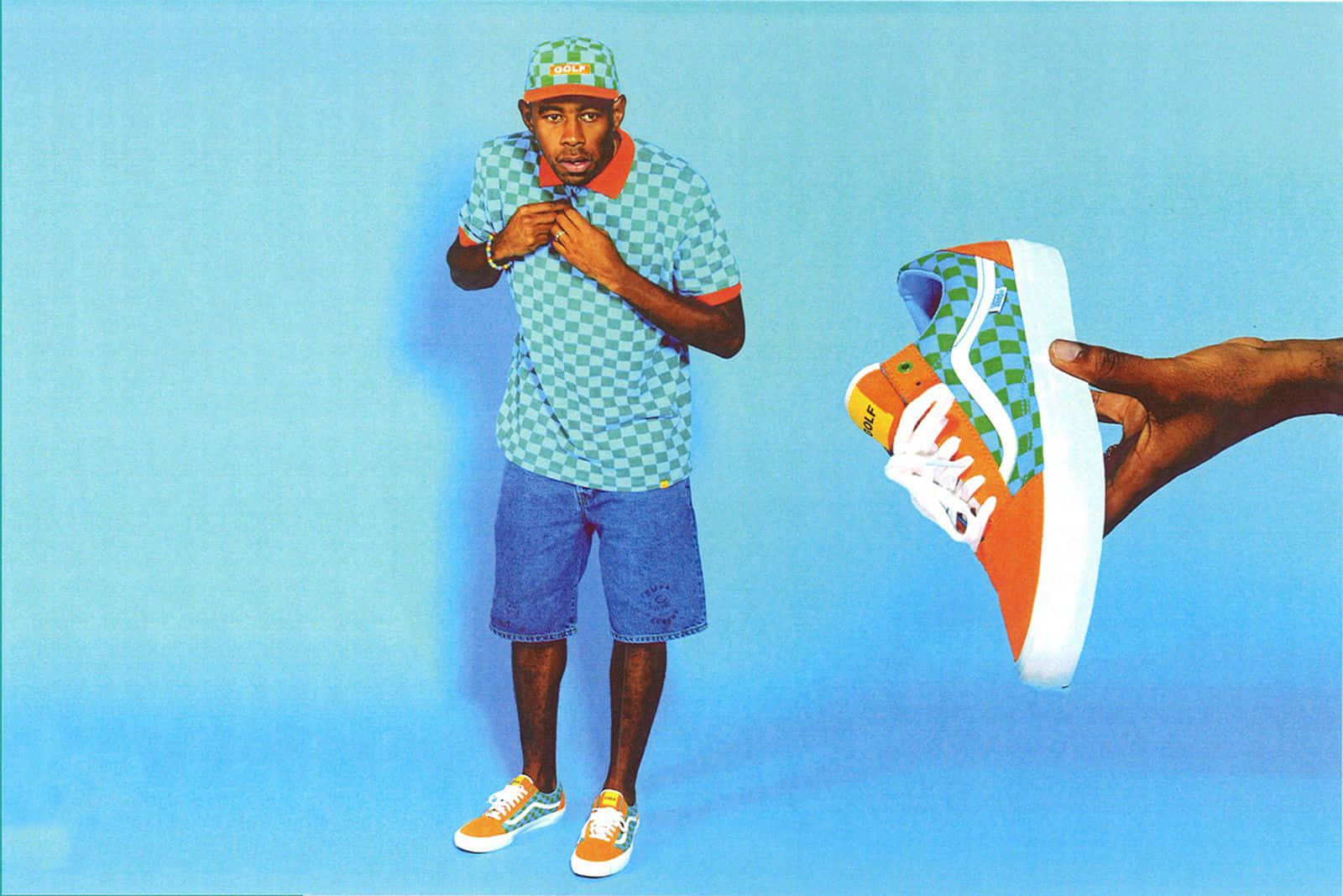 tyler the creator 桌布 壁紙 — 一名身穿格仔恤衫和帽子的男子站在藍色背景前，旁邊有一隻手拿著相配的運動鞋。