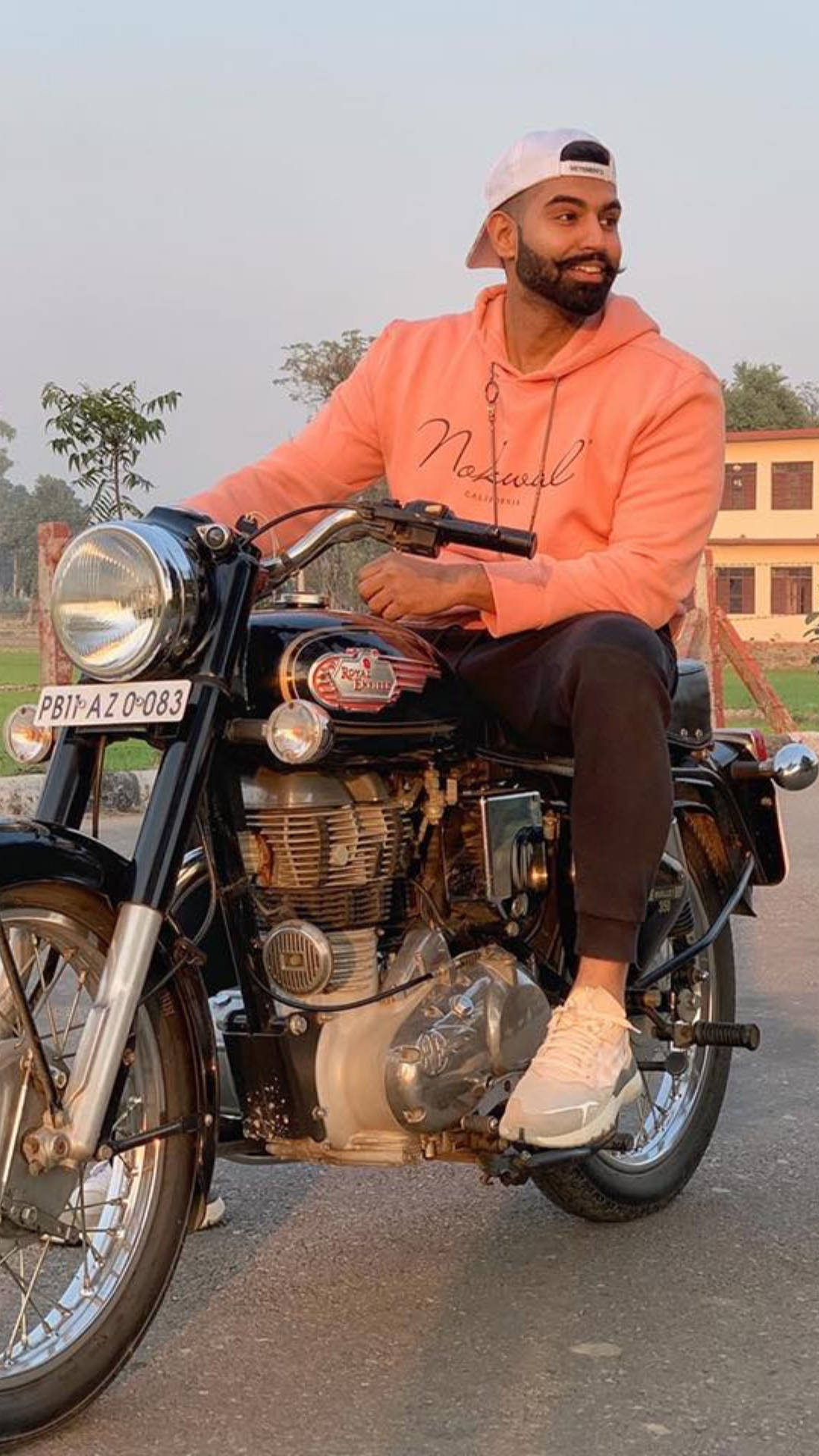 Indian 桌布 壁紙 — 一名身穿桃色連帽衫、戴白色帽子的男子微笑著坐在一輛帶有鍍鉻細節的經典黑色摩托車上，行駛在鋪砌的道路上。