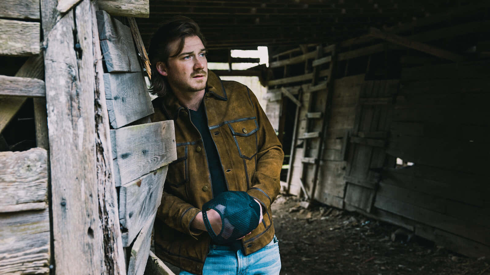 Morgan Wallen 桌布 壁紙 — 一名身穿棕色外套的男子靠喺殘舊穀倉入面嘅風化木板上，拎住一頂藍色帽。