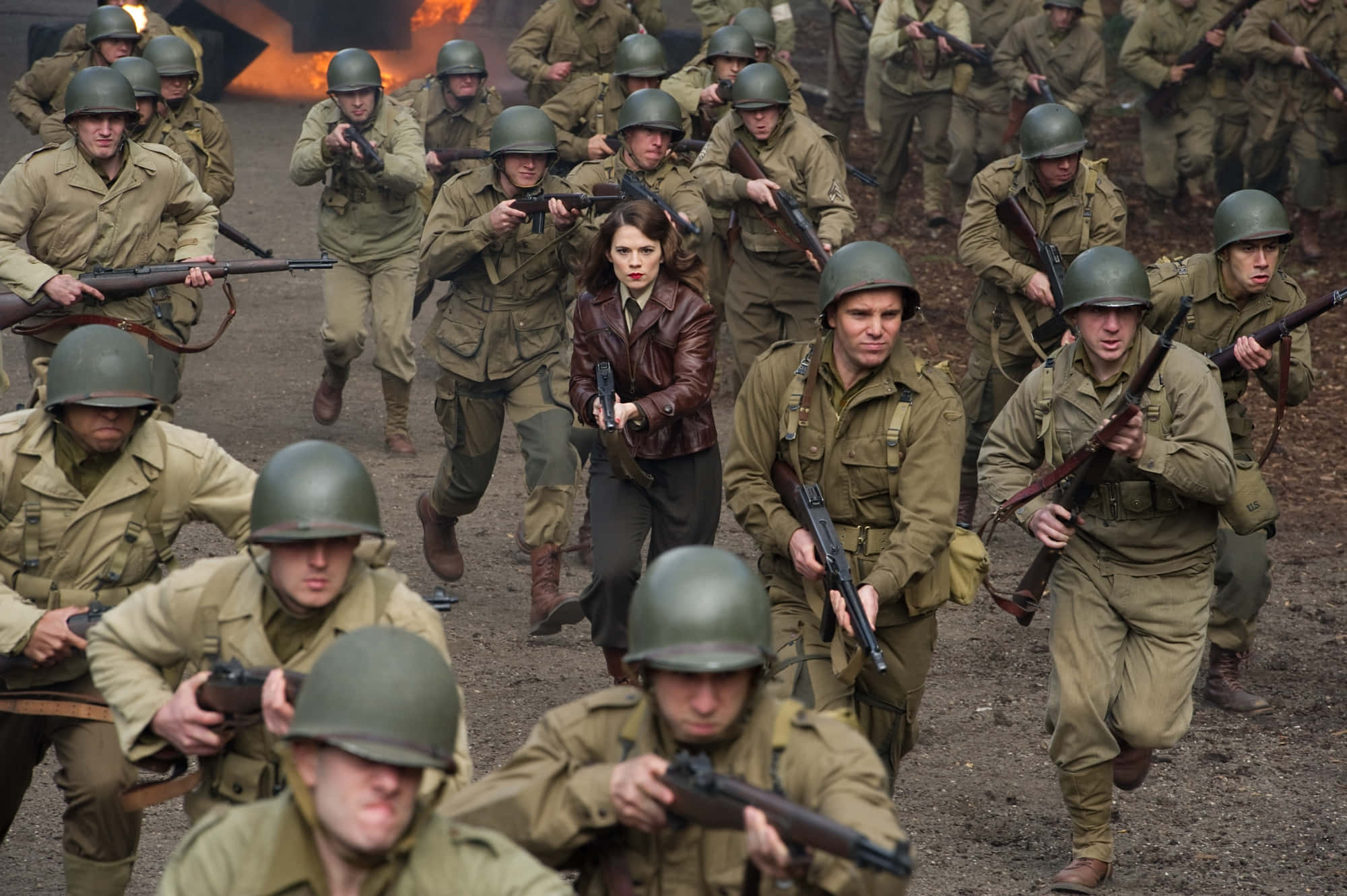 Peggy Carter 桌布 壁紙 — 一名身穿棕色皮夾克的女性在混亂的戰場上奔跑，周圍是穿著橄欖綠制服和頭盔的士兵。