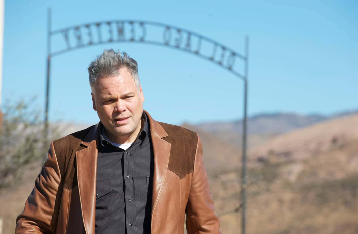 Vincent D'onofrio 桌布 壁紙 — 一名身穿棕色皮褸的男子，在晴朗的藍天下，站在一個風化的墓園大門前。