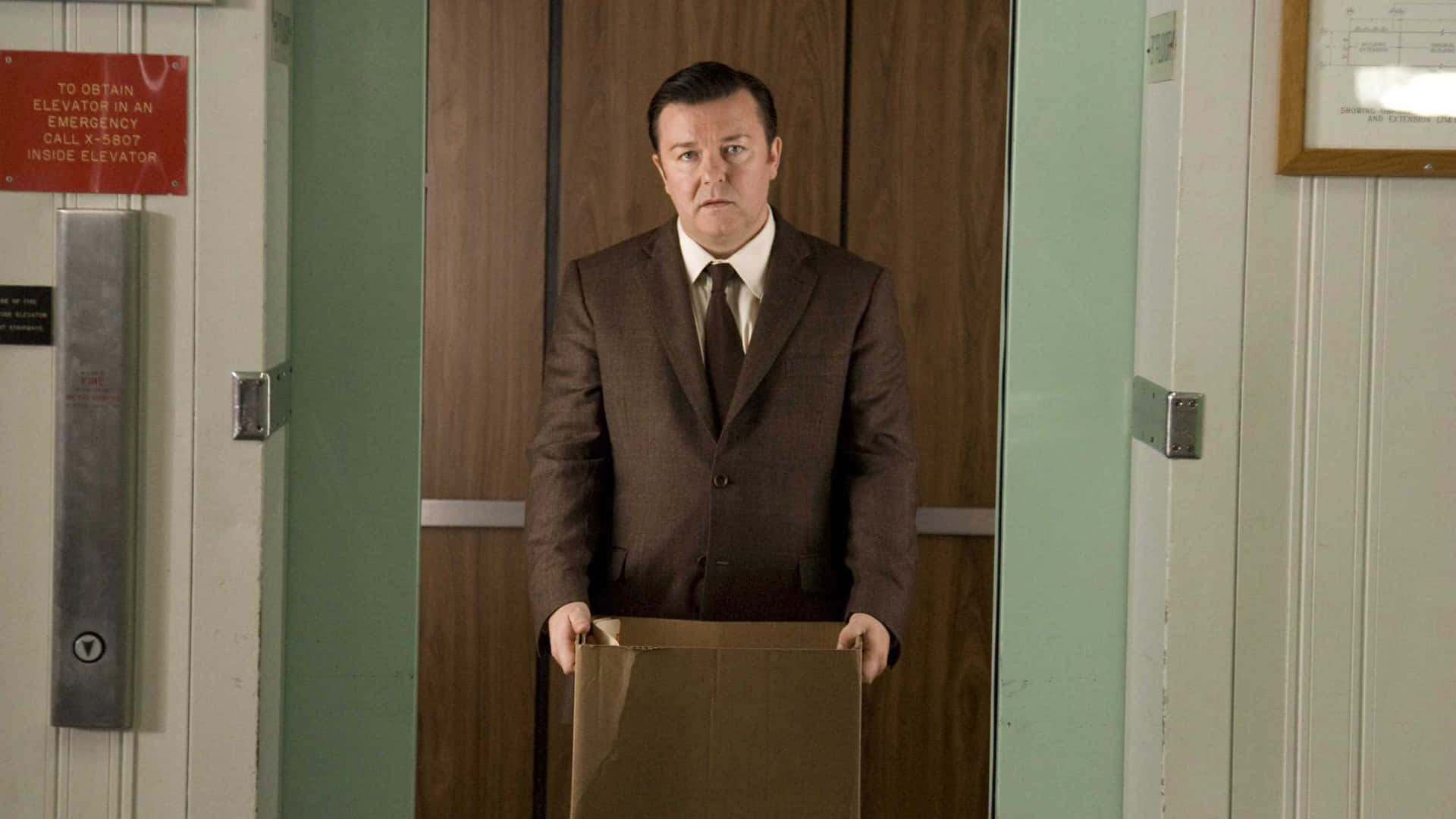 ricky gervais 桌布 壁紙 — 一名身穿棕色西裝的男子站在電梯大堂，手持紙皮箱，表情嚴肅。