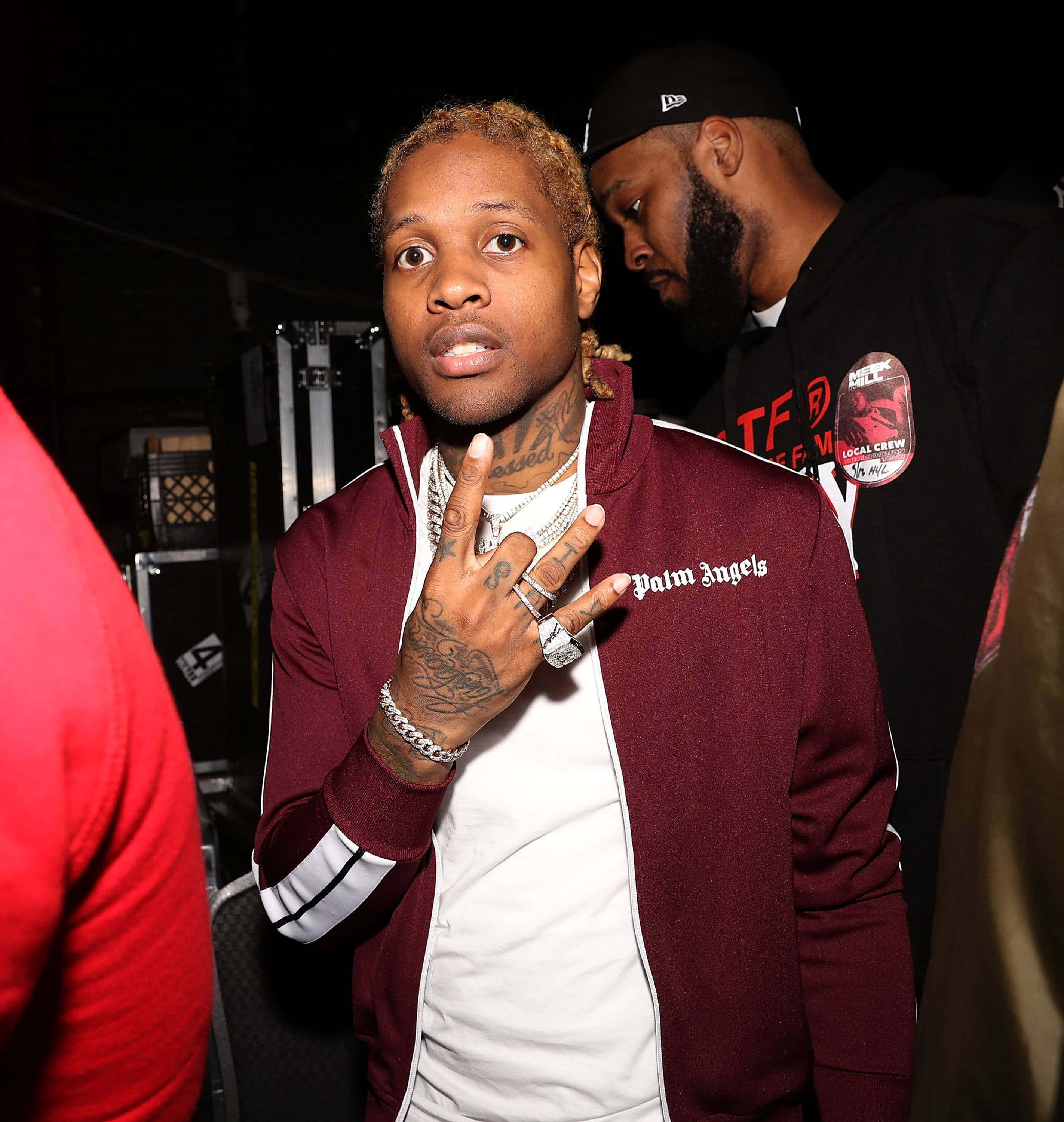 lil durk 桌布 壁紙 — 一名身穿棗紅色Palm Angels外套的男子在燈光昏暗的後台區域被人群包圍，做出和平手勢。