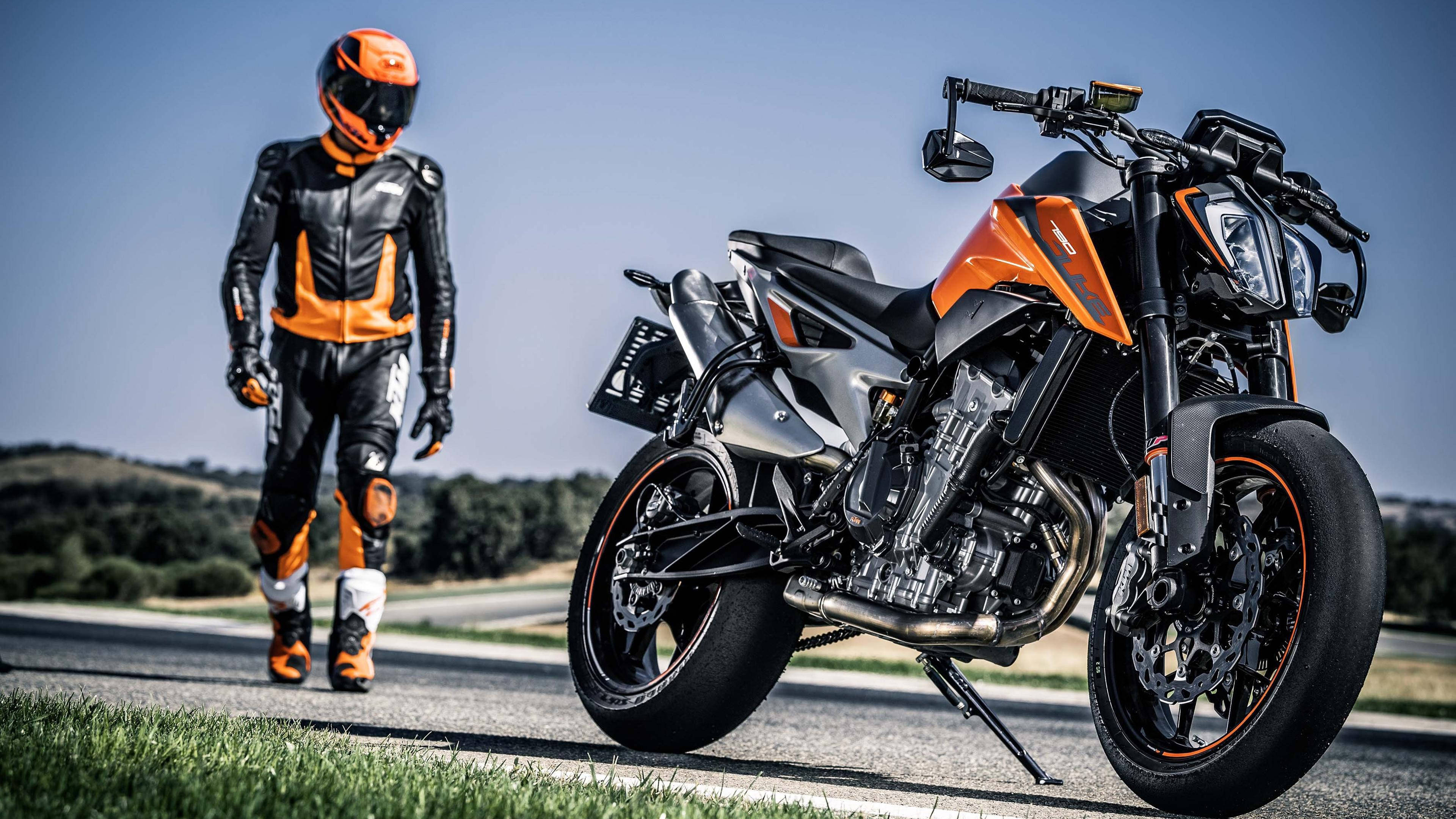 ktm bikes 桌布 壁紙 — 一名身穿橙色和黑色裝備的騎士，走在一輛停在陽光普照道路上的時尚橙色和黑色電單車旁邊。