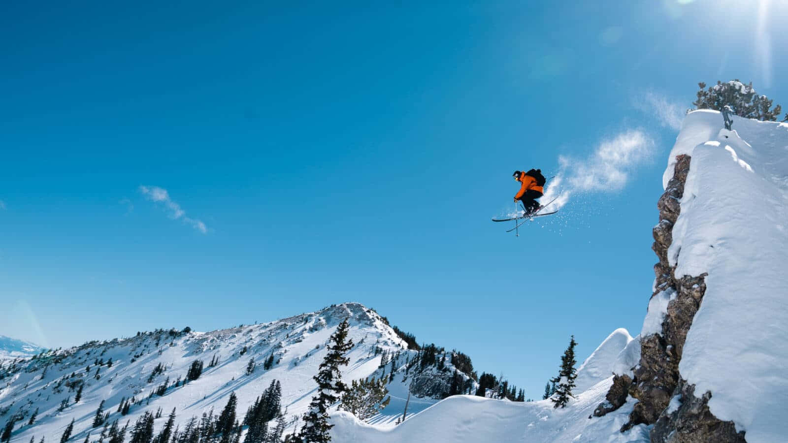 Skiing 桌布 壁紙 — 一名身穿橙色滑雪服的滑雪者從積雪懸崖上躍起，在清澈藍天下飛翔，下方是白雪覆蓋的山脈。