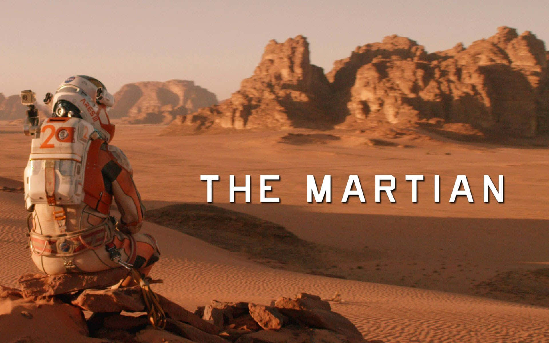 The Martian 桌布 壁紙 — 一名身穿橙色裝備的太空人跪在岩石遍佈的火星沙漠上，凝視著朦朧天空下遠處的紅色岩層。