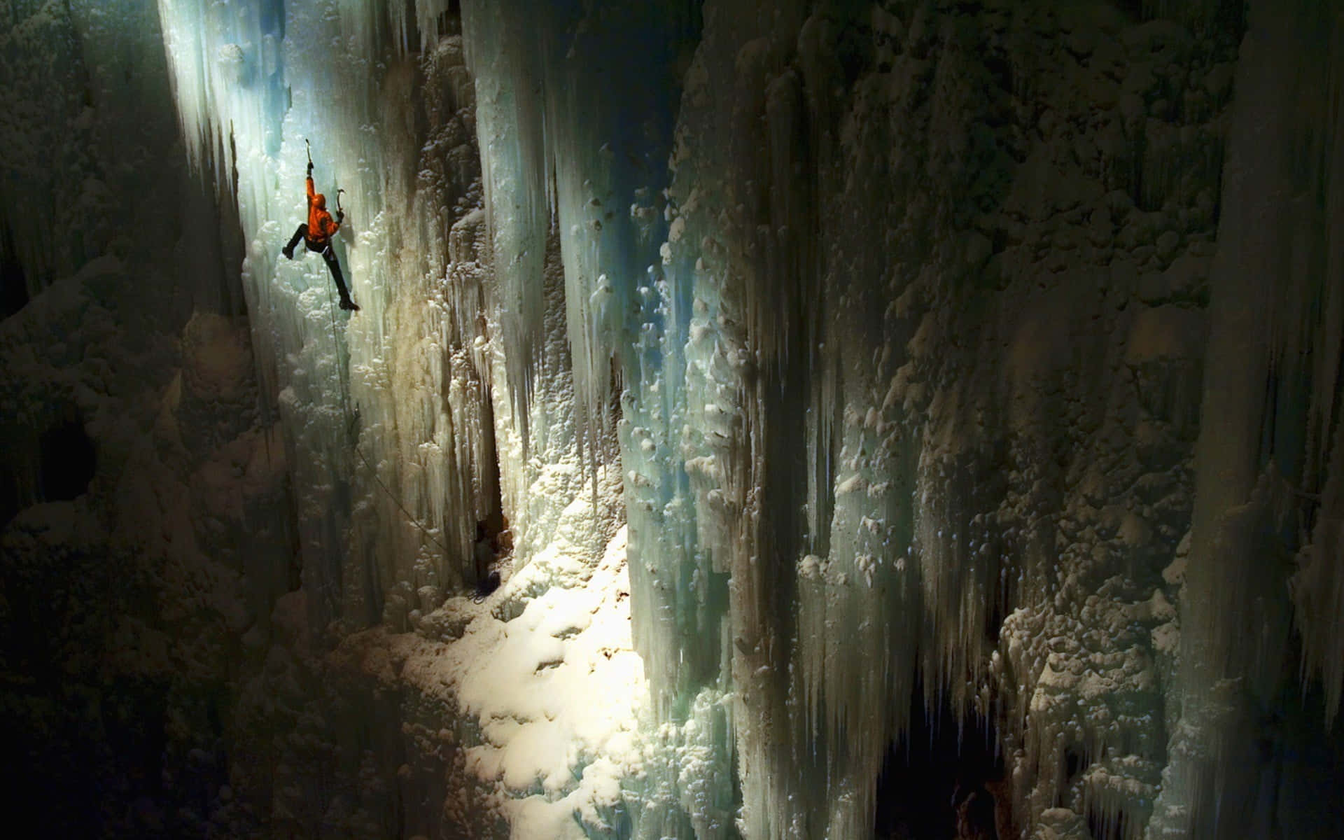 Ice Climbing 桌布 壁紙 — 一名身穿橙色裝備的攀岩者正在一個閃爍藍光的冰洞內，攀爬一座高聳的冰壁，洞內懸掛著冰柱。