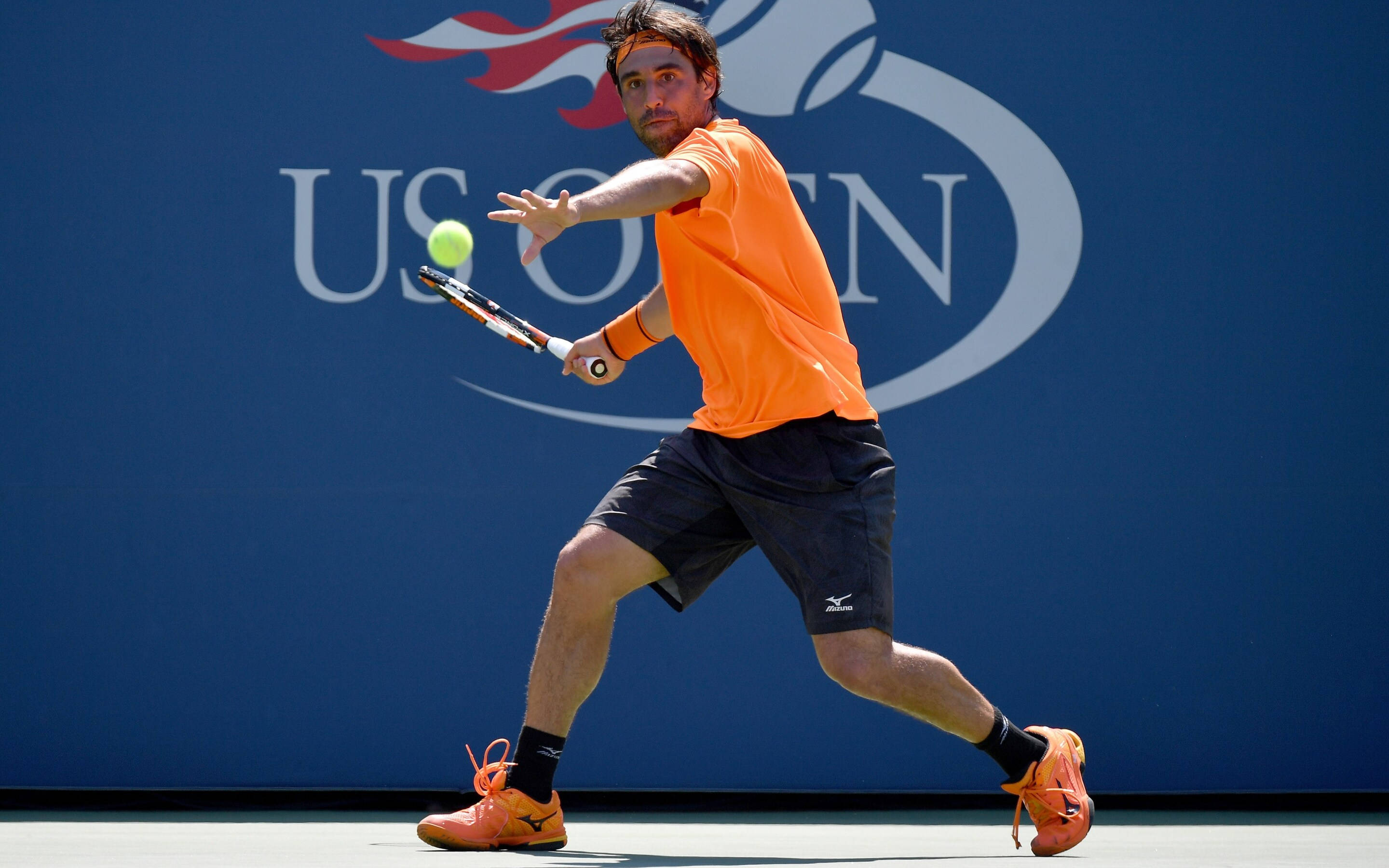 Marcos Baghdatis Playing Tennis 壁紙 — 一名身穿橙色裝備的男網球手在藍色美國公開賽背景下揮拍擊球。
