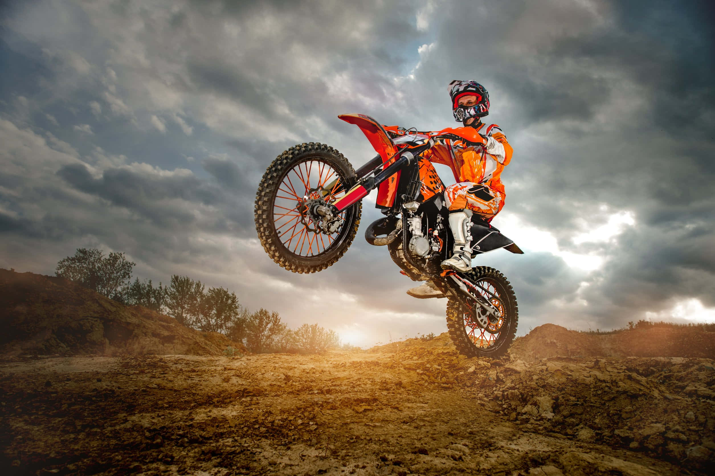 Dirtbike 桌布 壁紙 — 一名身穿橙色裝備的越野電單車手，在日落時分戲劇性的多雲天空背景下，騎著紅色電單車在空中飛翔。