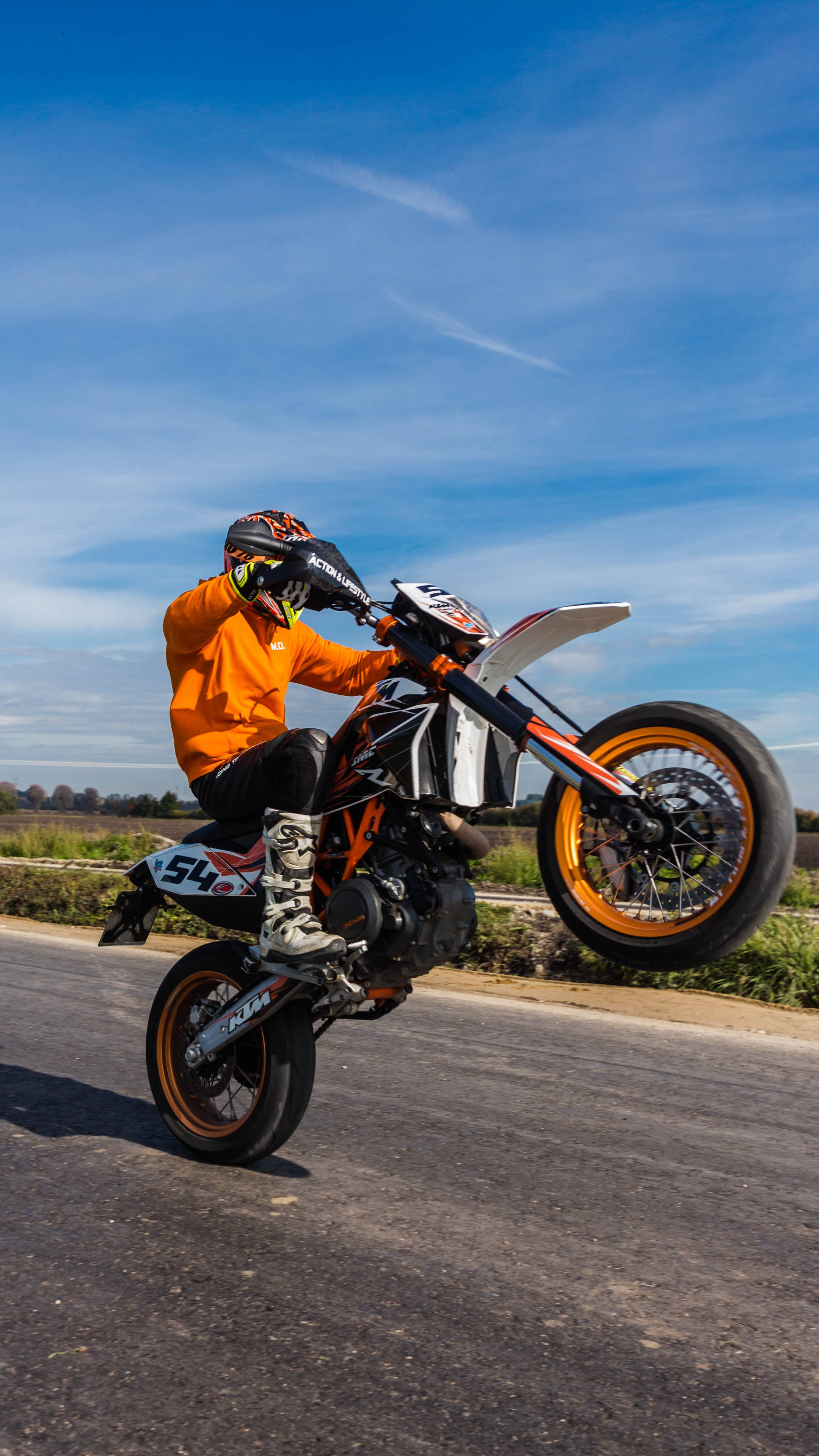 ktm rc 200 桌布 壁紙 — 一名身穿橙色裝備的騎手在陽光明媚的道路上，駕駛橙色和白色的KTM越野電單車表演翹前輪。