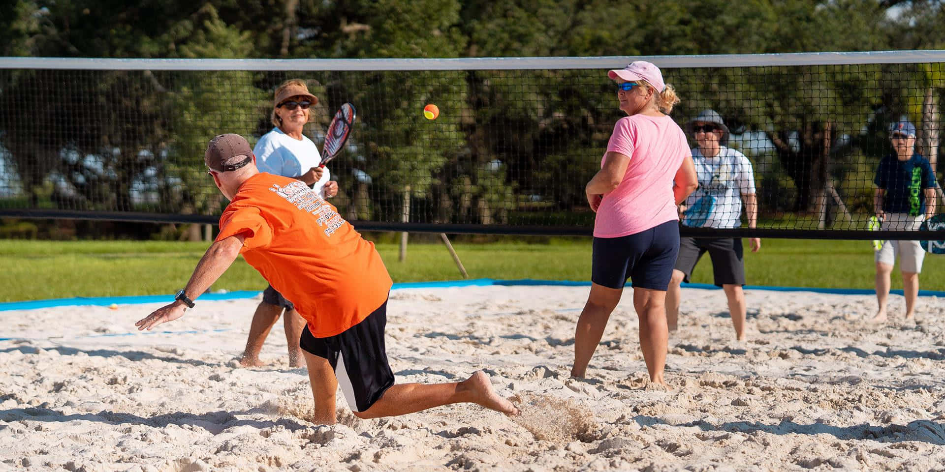 Beach Tennis 桌布 壁紙 — 一名身穿橙色襯衫的男子在沙地球場上飛身撲向排球，陽光普照的沙灘比賽中，觀眾在旁觀看。