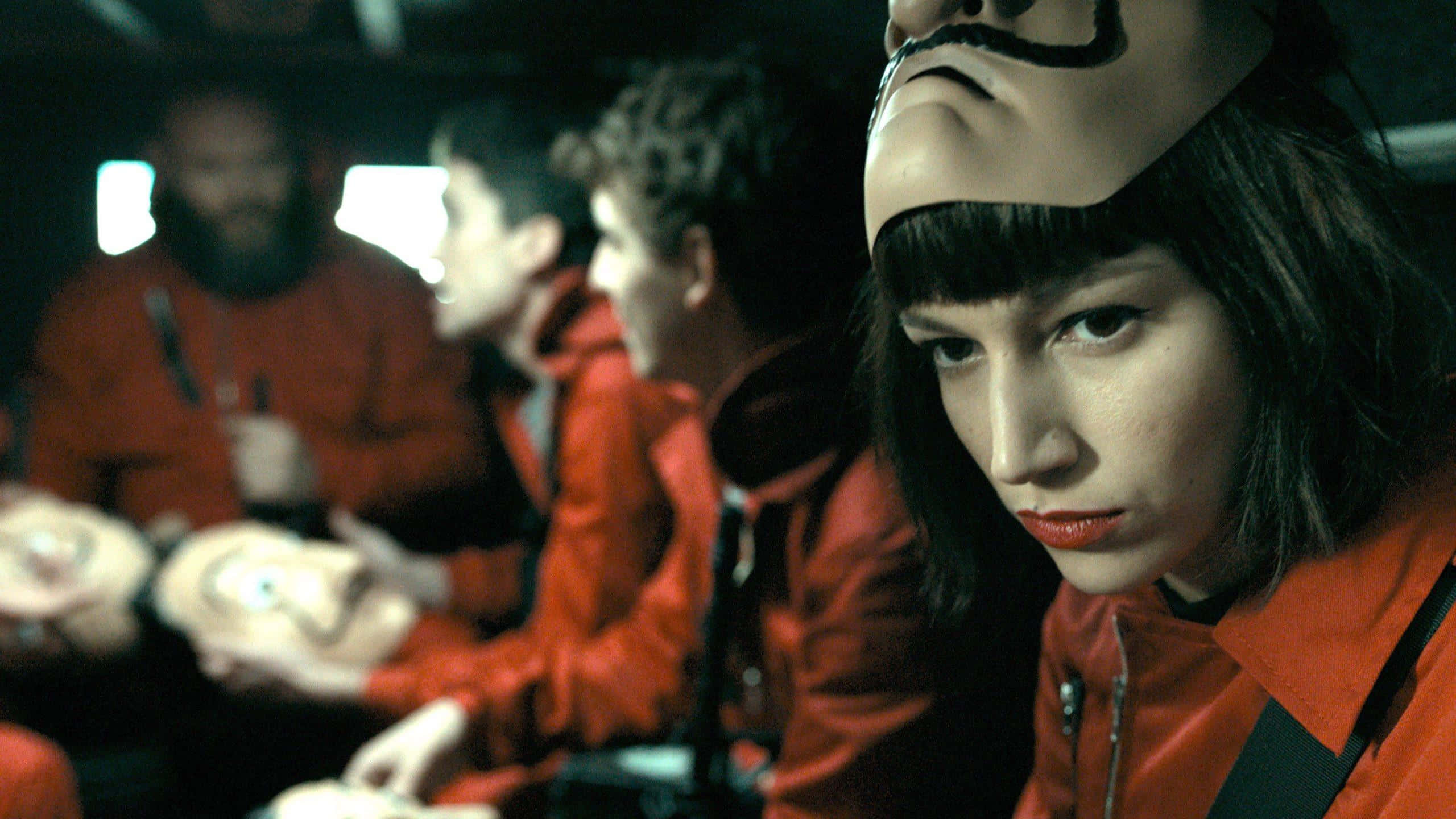 Money Heist 桌布 壁紙 — 一名身穿橙色連身衣和白色口罩的女子專注地凝視，而其他穿著類似服裝的人坐在她身後一個光線昏暗的房間裡。