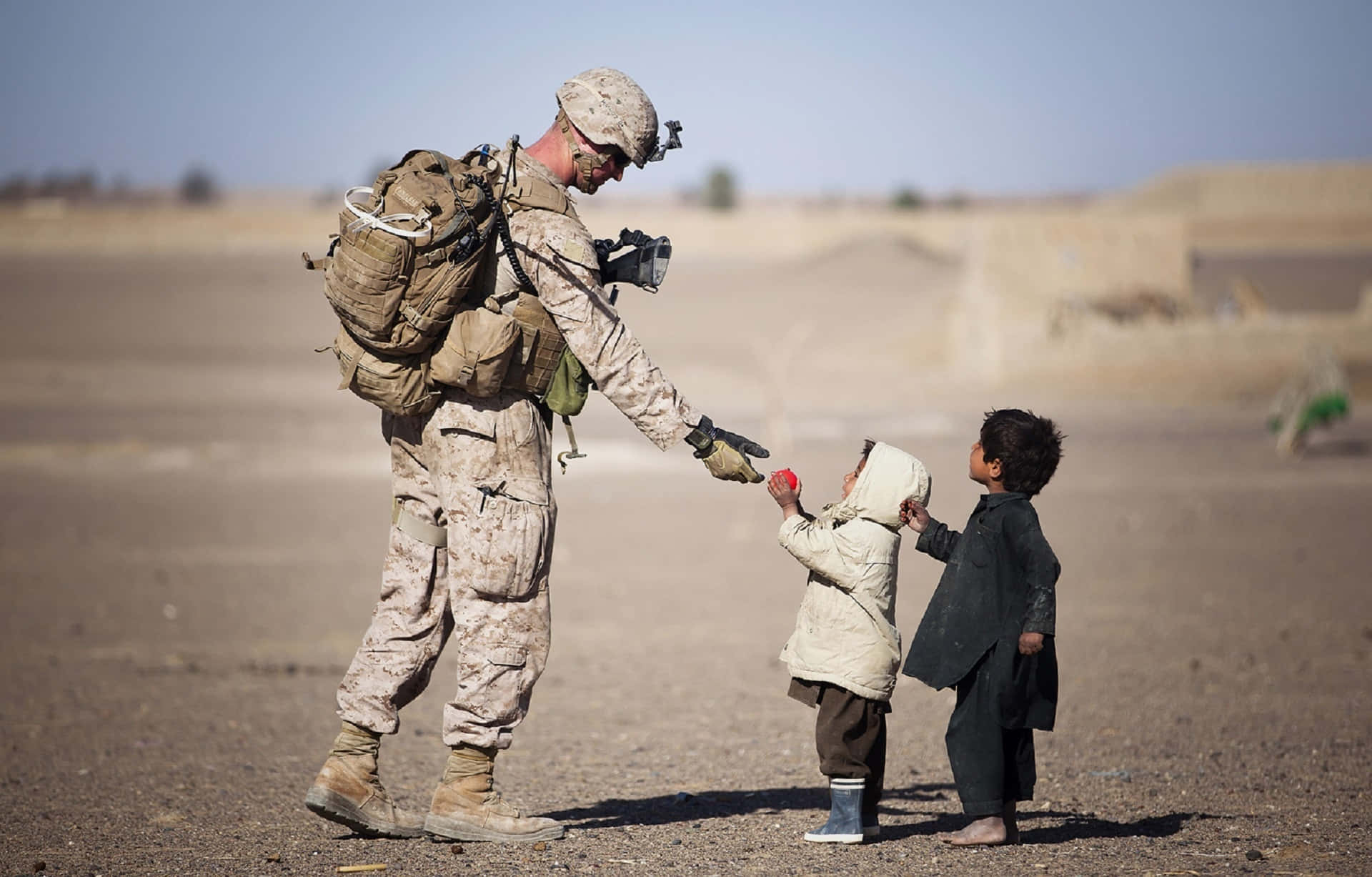 children 桌布 壁紙 — 一名身穿沙漠迷彩服的士兵在烈日下，於塵土飛揚的貧瘠景觀中，與兩個赤腳的孩子分享一個紅蘋果。