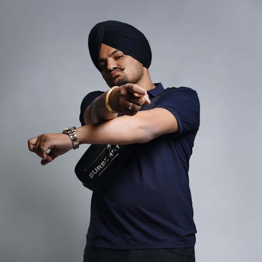 Sidhu Moose Wala 桌布 壁紙 — 一名身穿海軍藍頭巾和Polo衫的男子自信地指著手錶，背景是純灰色。