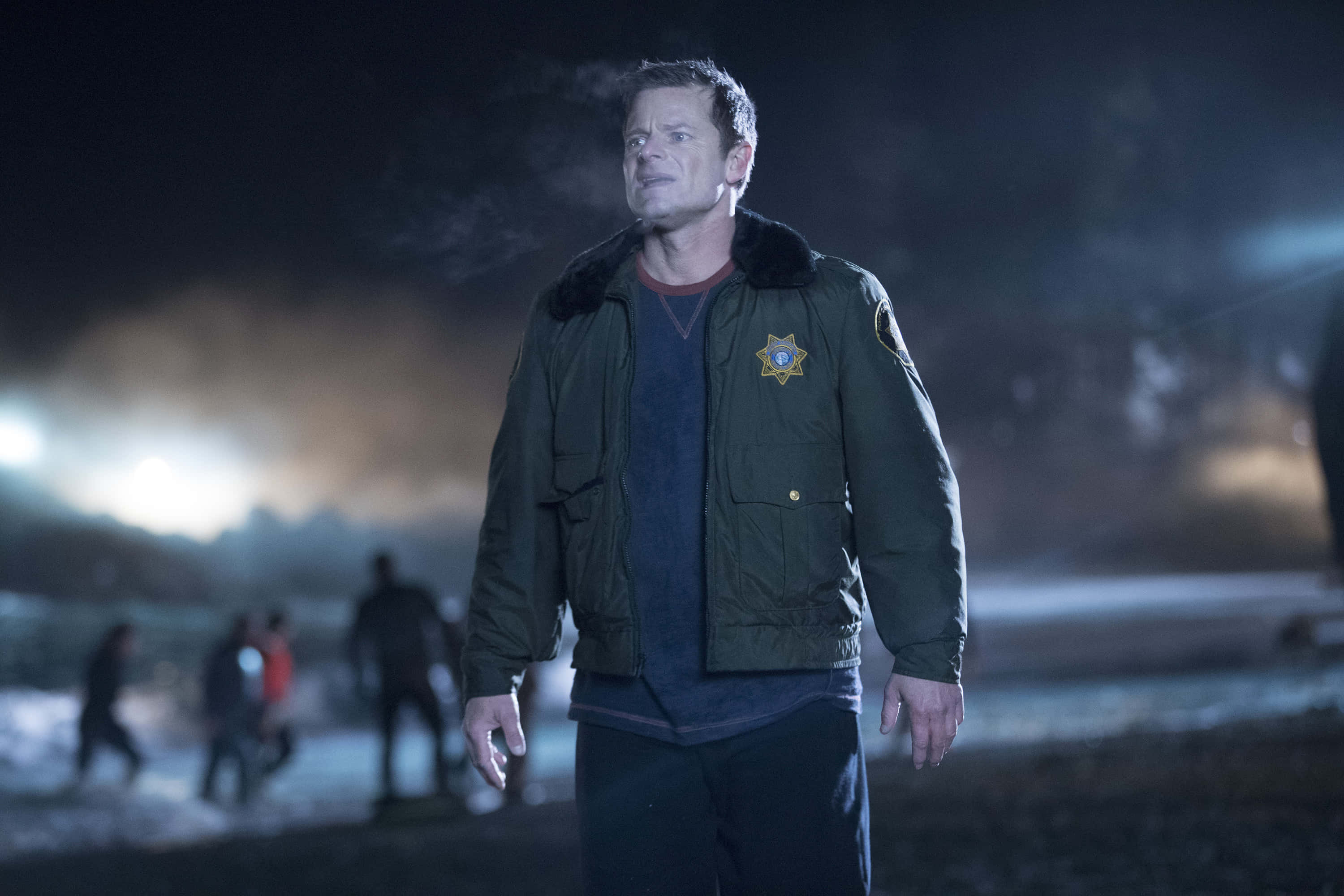 Steve Zahn 桌布 壁紙 — 一名身穿深綠色外套的警長在夜間保持警覺，背景中模糊的人影在刺眼的人造燈光下移動。