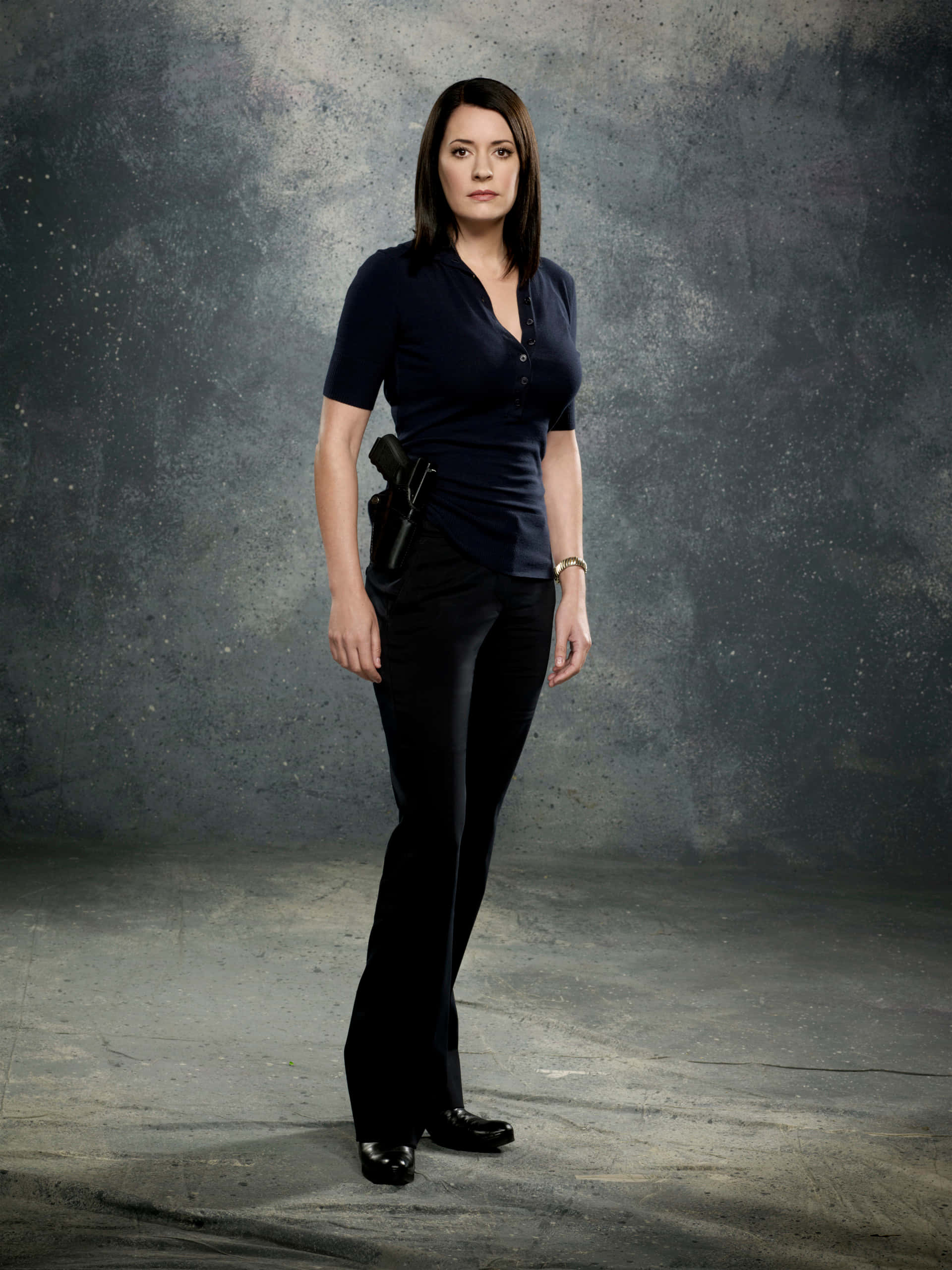 paget brewster 桌布 壁紙 — 一名身穿深色制服嘅女人企喺粗糙紋理嘅灰色背景前，腰間槍套插住手槍，姿勢穩健。