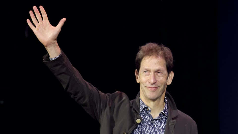 Tim Blake Nelson 桌布 壁紙 — 一名身穿深色外套的男子在明亮的舞台燈光下，對著黑色背景愉快地揮手。