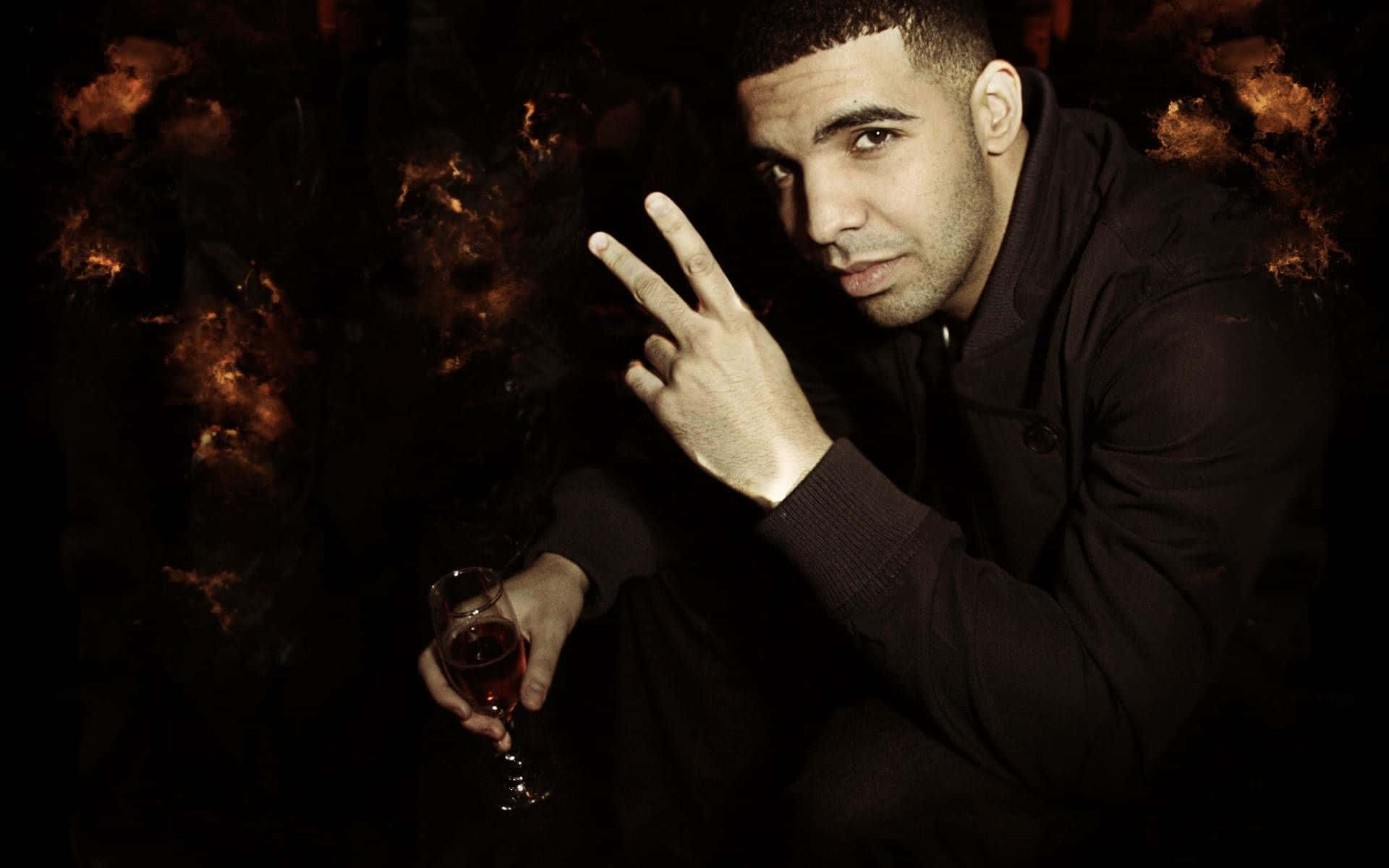 Drake 桌布 壁紙 — 一名身穿深色外套的男子在煙霧瀰漫的火光背景下，手持一杯紅酒做出和平手勢。