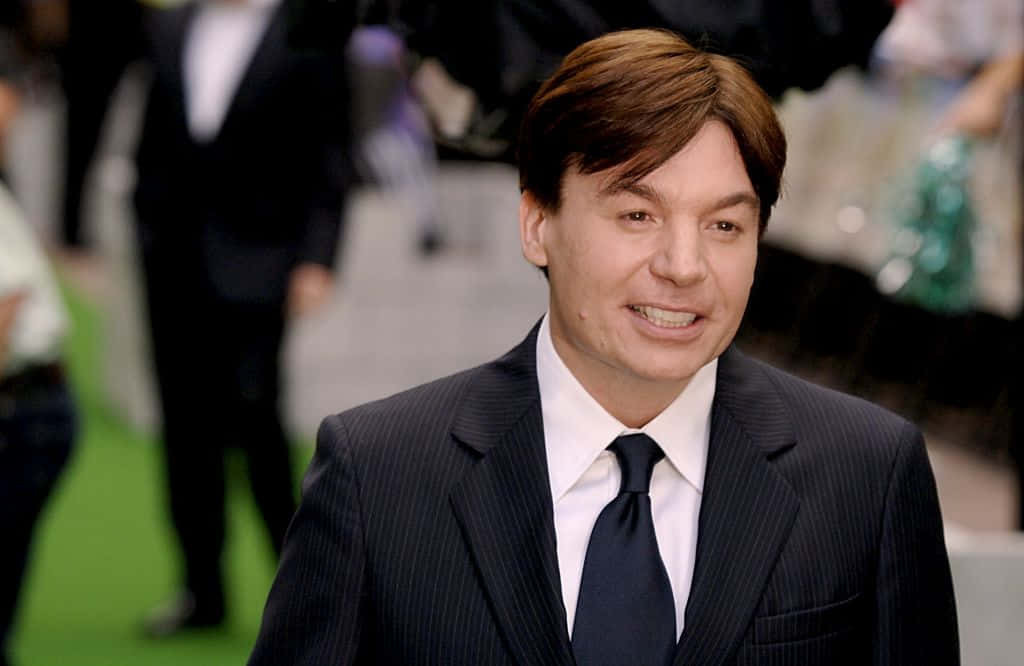 Mike Myers 桌布 壁紙 — 一名身穿深色細條紋西裝和領帶的男子，在綠色地毯上對著鏡頭溫暖微笑，身後有模糊的人群。
