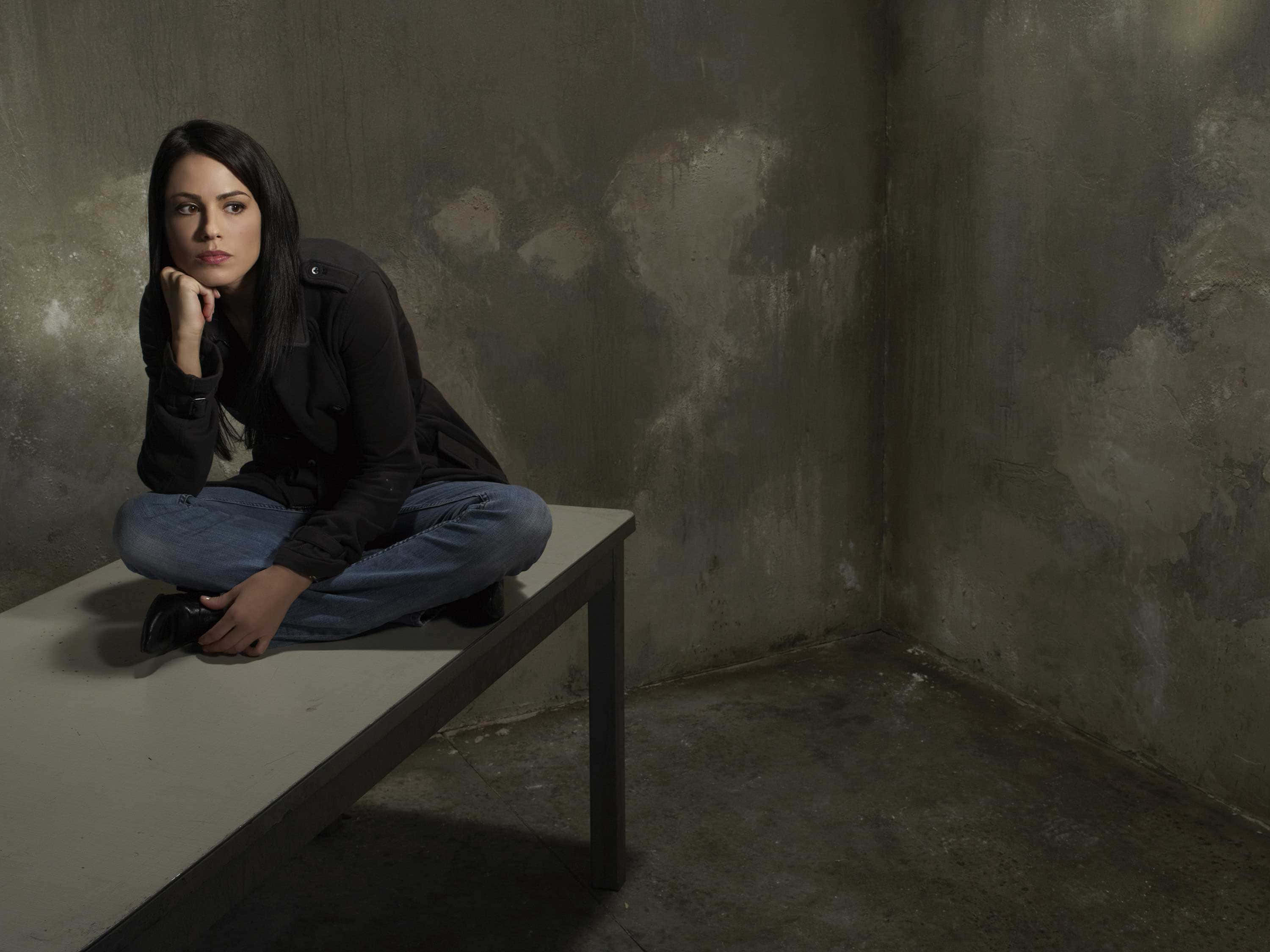 Michelle Borth 桌布 壁紙 — 一名身穿深色衣服的女子在光線昏暗的房間裏，靠著風化的混凝土牆，若有所思地坐在長椅上。
