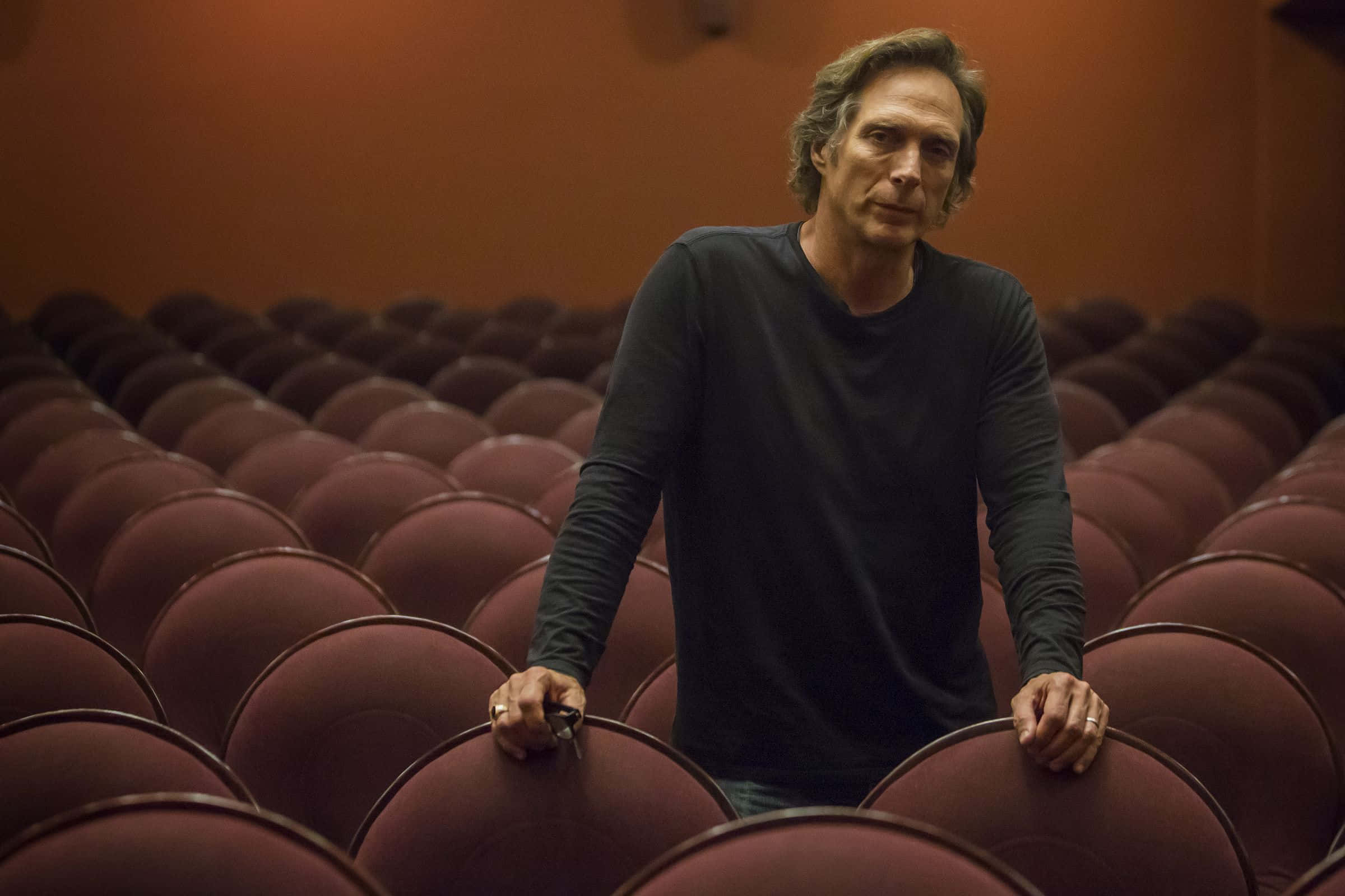 William Fichtner 桌布 壁紙 — 一名身穿深色襯衫的男子，在溫暖的環境燈光下，莊嚴地站在一排排空蕩蕩的紅色劇院座位之間。