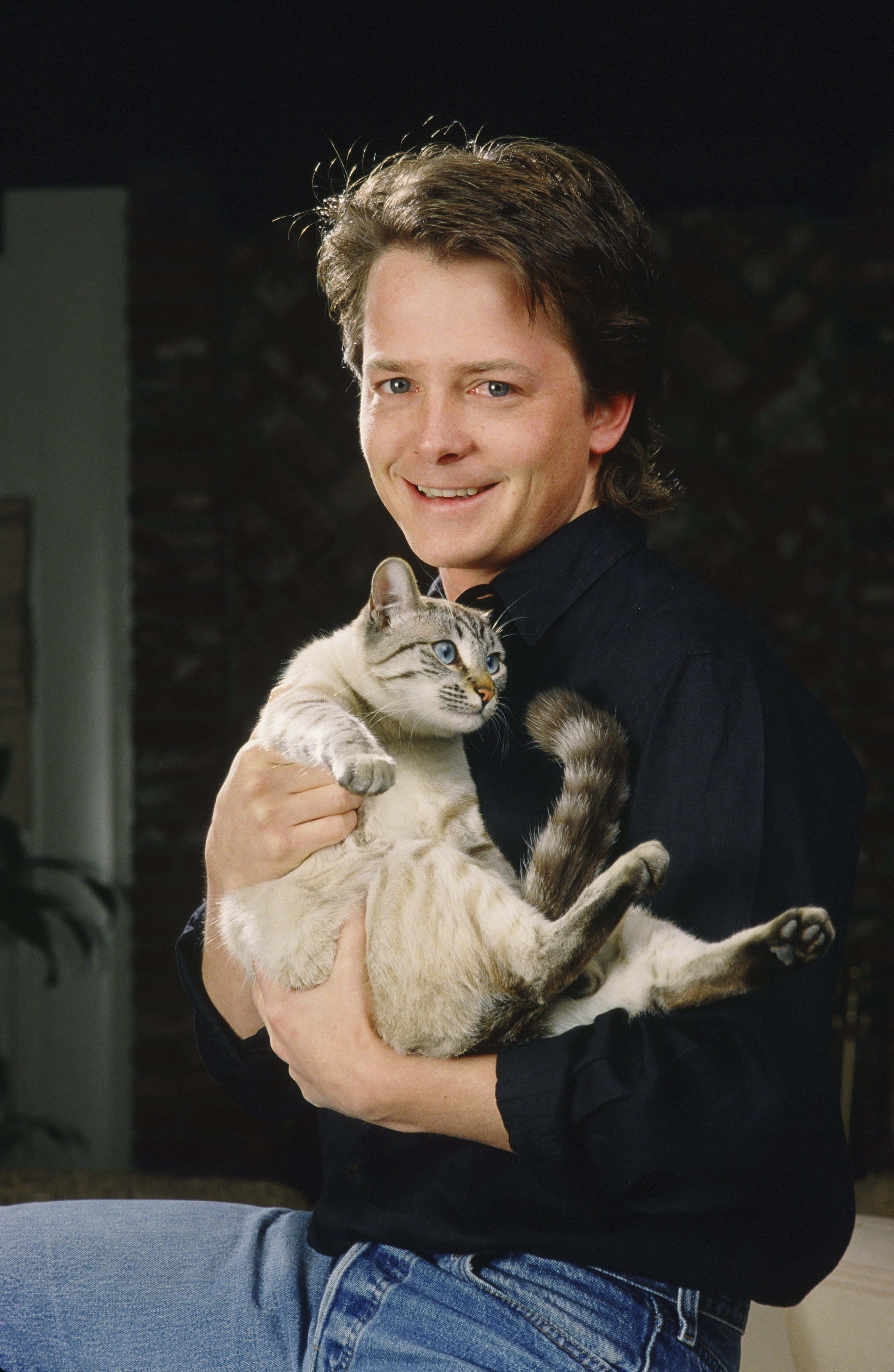 michael j fox 桌布 壁紙 — 一名身穿深色襯衫的男子在昏暗的室內背景下，溫暖地微笑著抱著一隻藍眼睛的淺灰色虎斑貓。