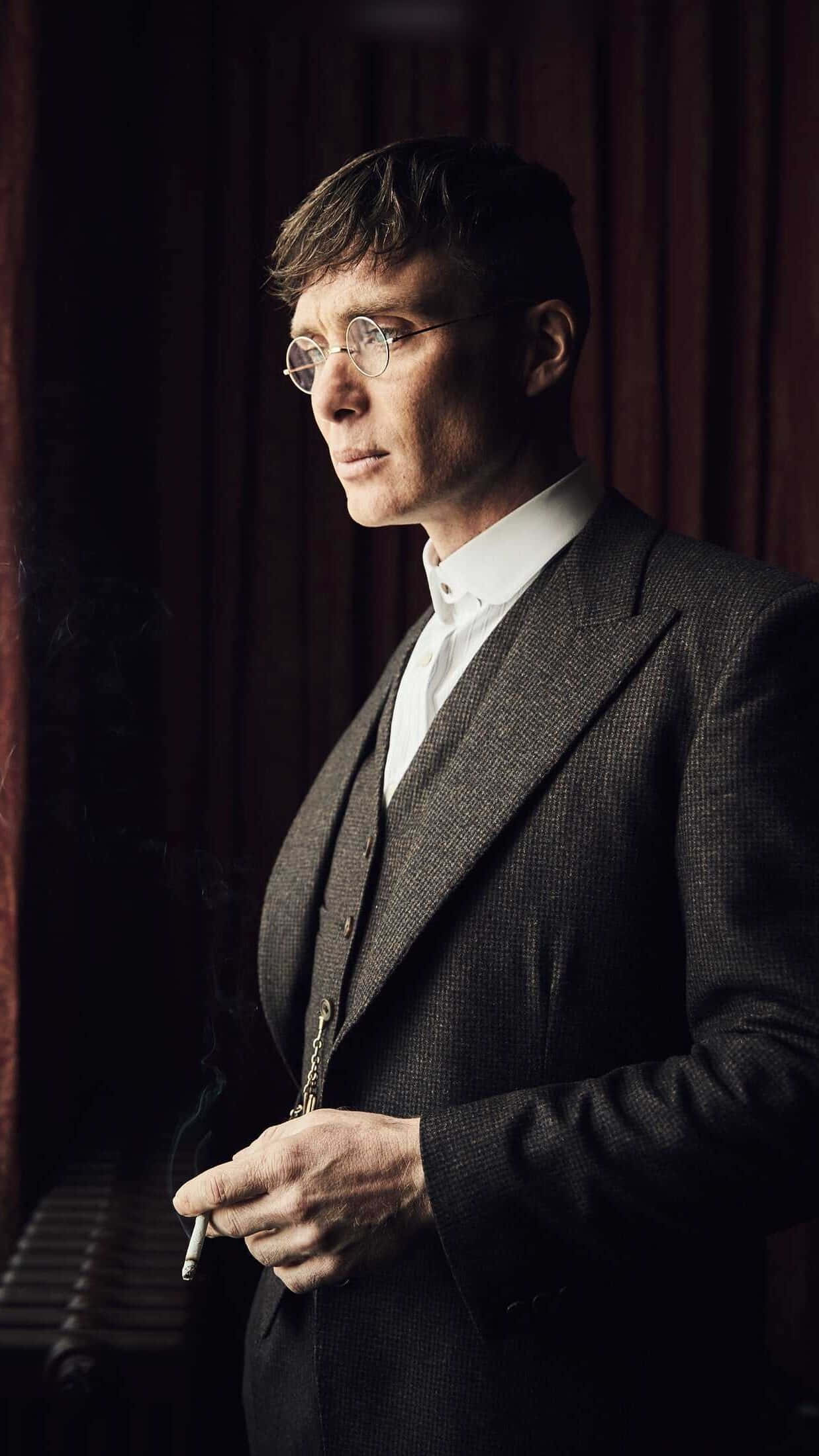 Peaky Blinders 桌布 壁紙 — 一名身穿深色西裝、戴圓框眼鏡的男子，在厚重的紅色窗簾背景下吸煙。