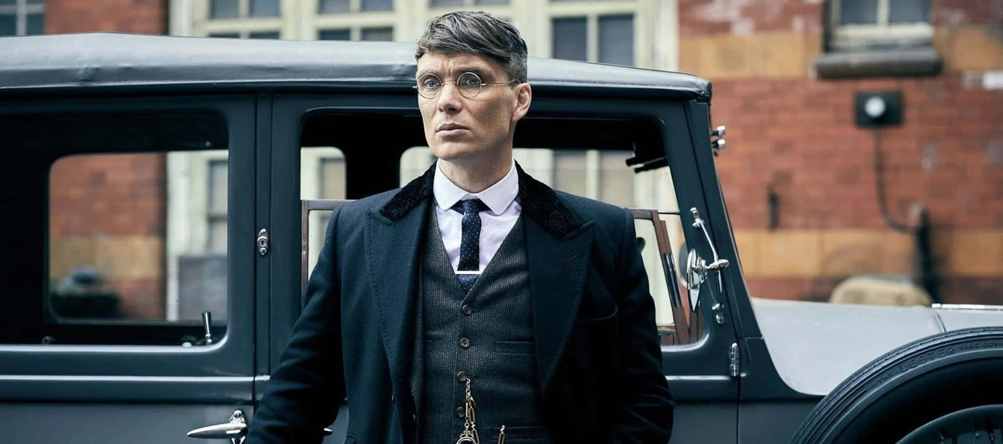 Peaky Blinders 桌布 壁紙 — 一名身穿深色西裝、戴眼鏡的男子站在磚砌建築旁，旁邊是一輛復古黑色汽車。