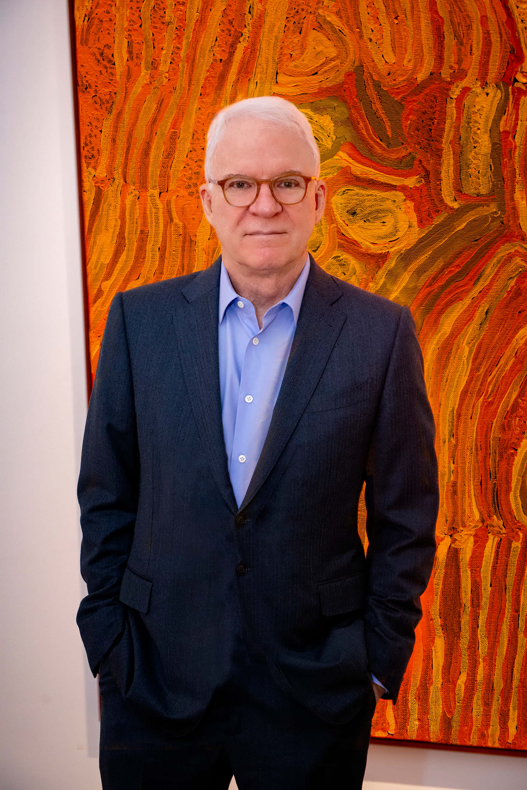 steve martin 桌布 壁紙 — 一名身穿深色西裝、戴著眼鏡的男子自信地雙手插袋，站在一幅充滿活力的抽象畫前，畫中有旋轉的橙色和黃色筆觸。