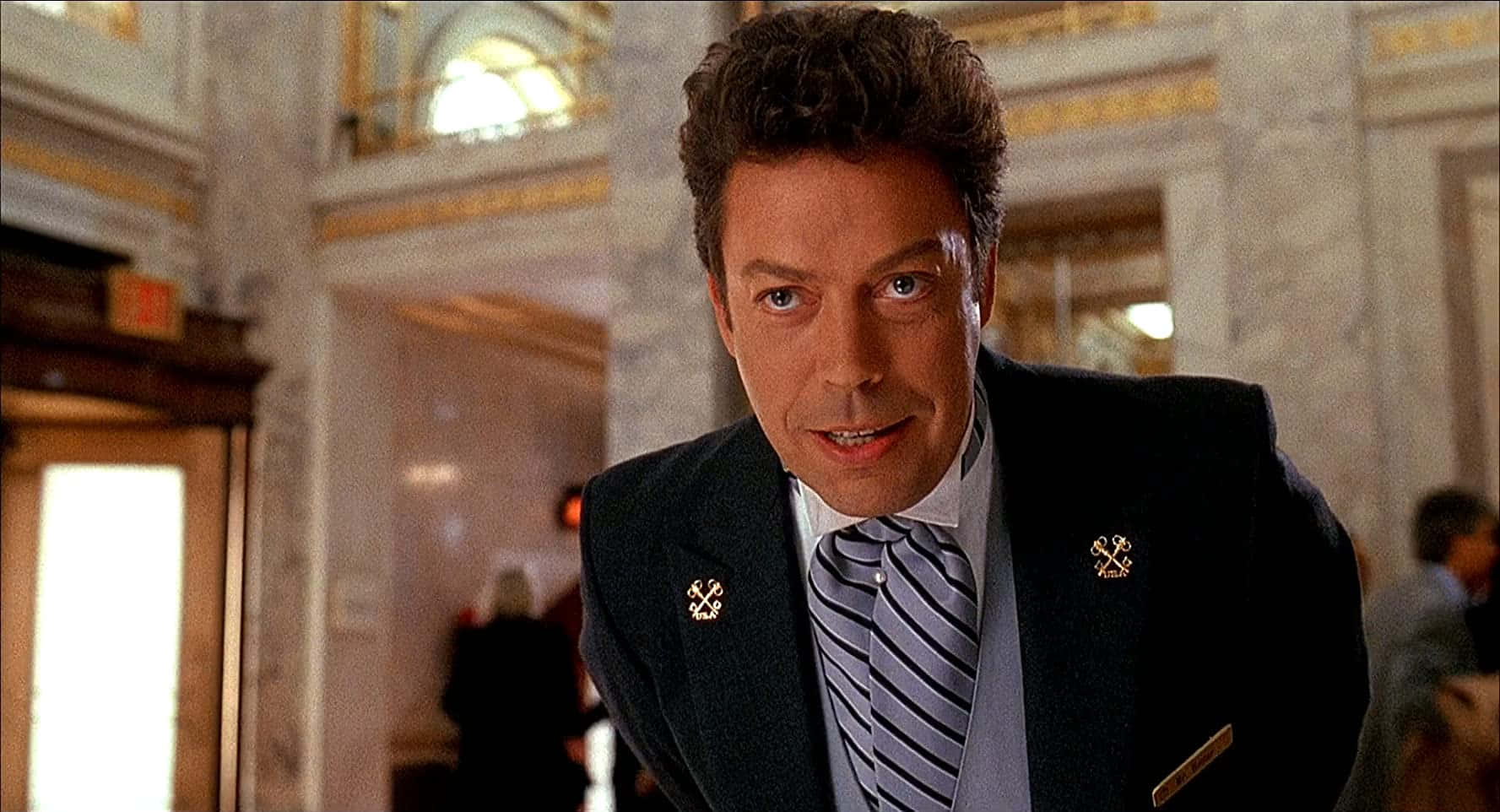 Tim Curry 桌布 壁紙 — 一名身穿深色西裝、戴著金色領帶夾的條紋領帶的男子，在宏偉的大理石走廊中專注地向前看。
