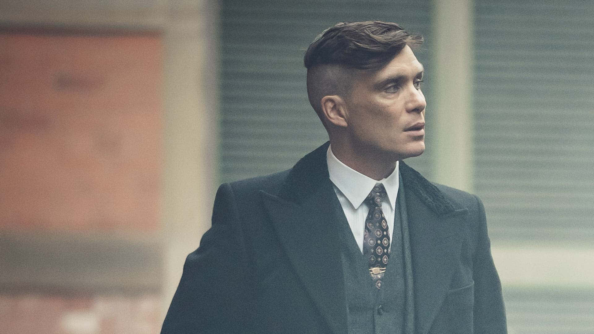 Peaky Blinders 桌布 壁紙 — 一名身穿深色西裝及圖案領帶的男子站在霧氣瀰漫的都市街道上，背景是磚造建築。