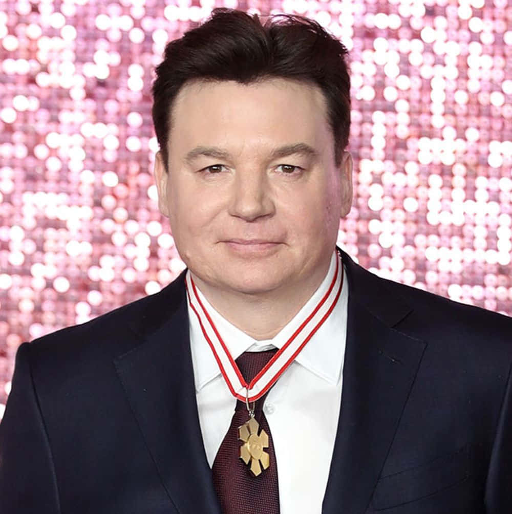 Mike Myers 桌布 壁紙 — 一名身穿深色西裝及領帶的男子，佩戴著紅白絲帶上的金牌，背景是閃爍的粉紅色。