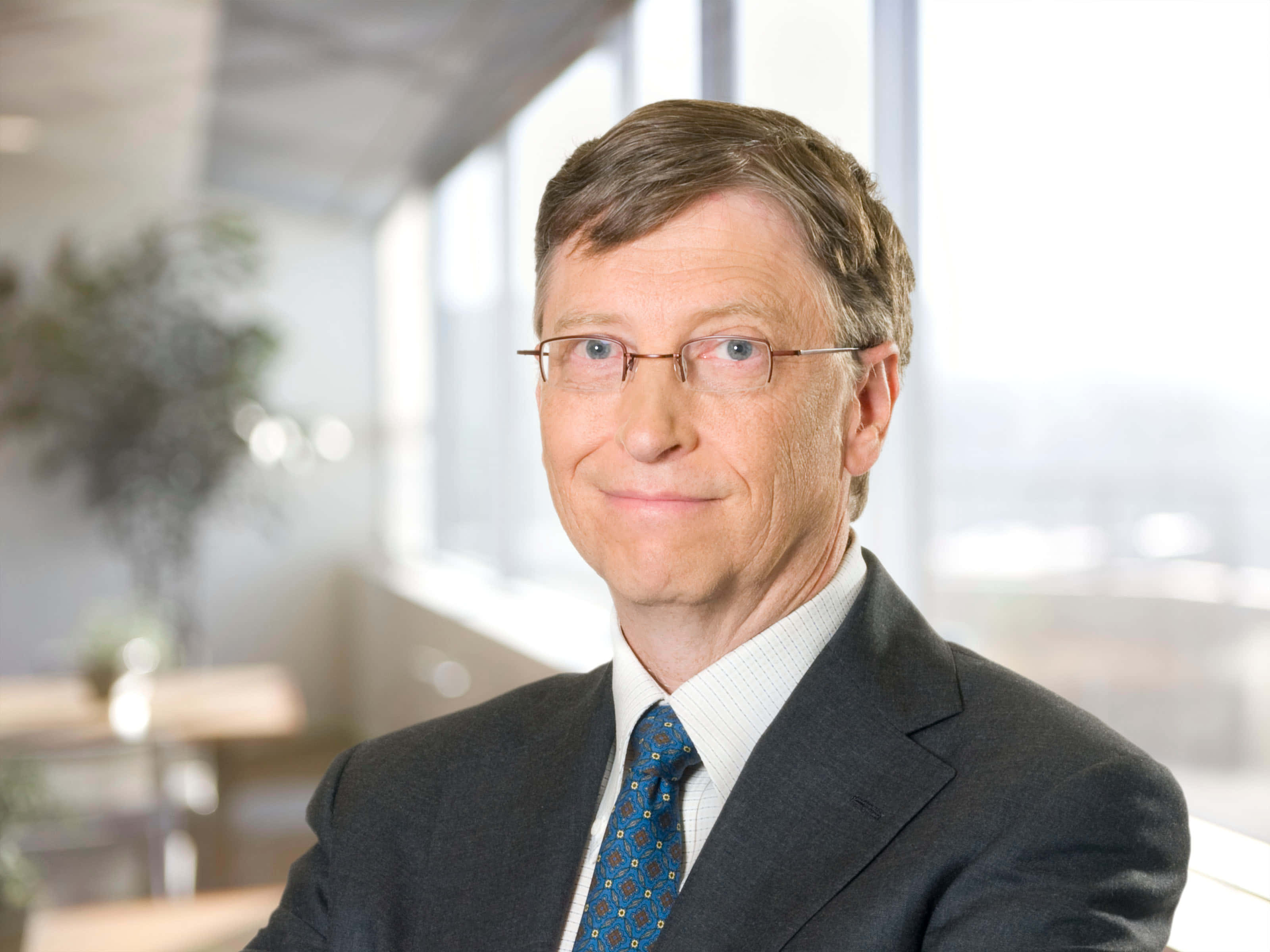 Bill Gates 桌布 壁紙 — 一名身穿深色西裝和圖案領帶的男子，坐在明亮現代、有大窗戶的辦公室裡，溫柔地微笑。