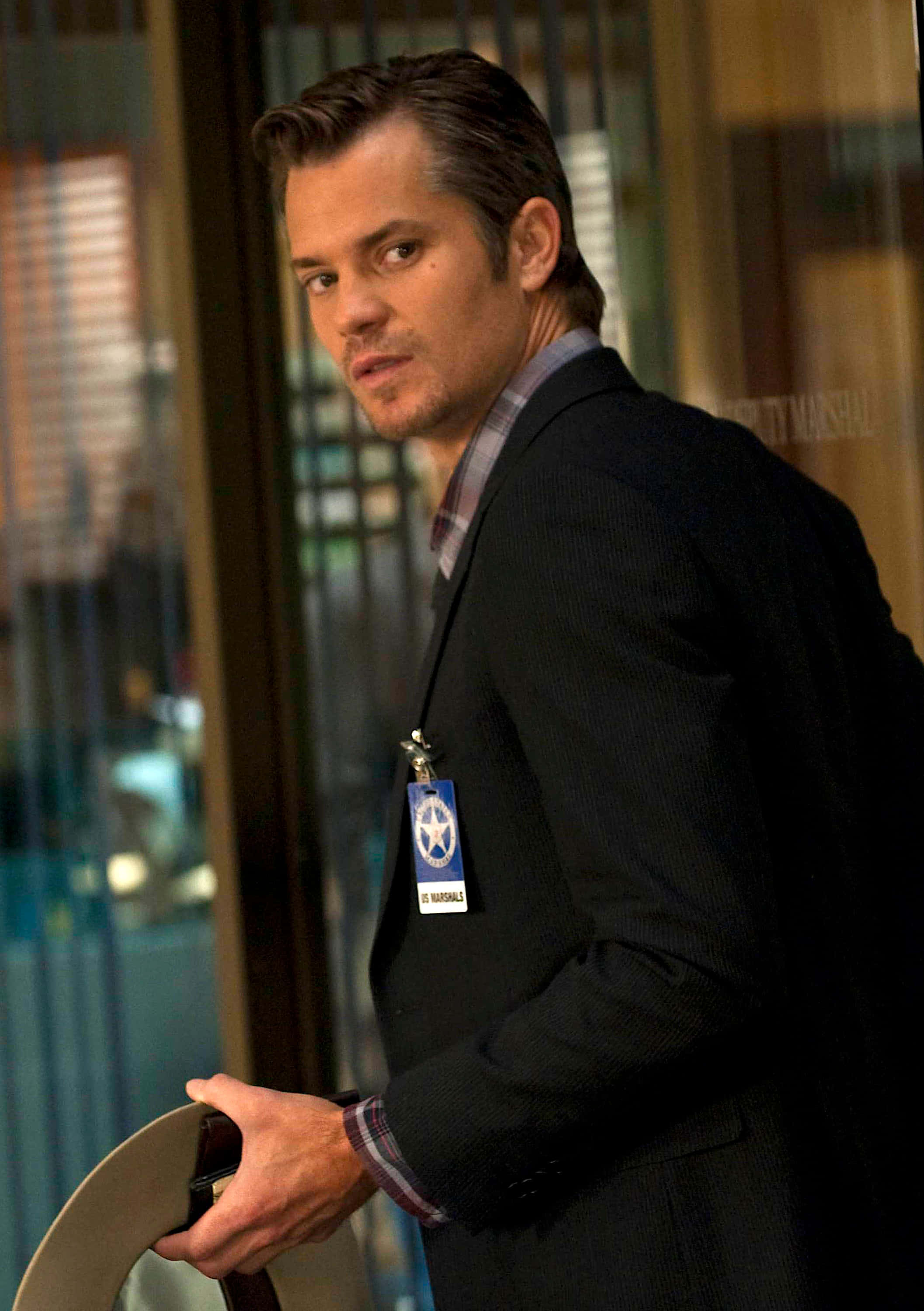 timothy olyphant 桌布 壁紙 — 一名身穿深色西裝和徽章的保安員站在玻璃門口，手持帽子，神情嚴肅。