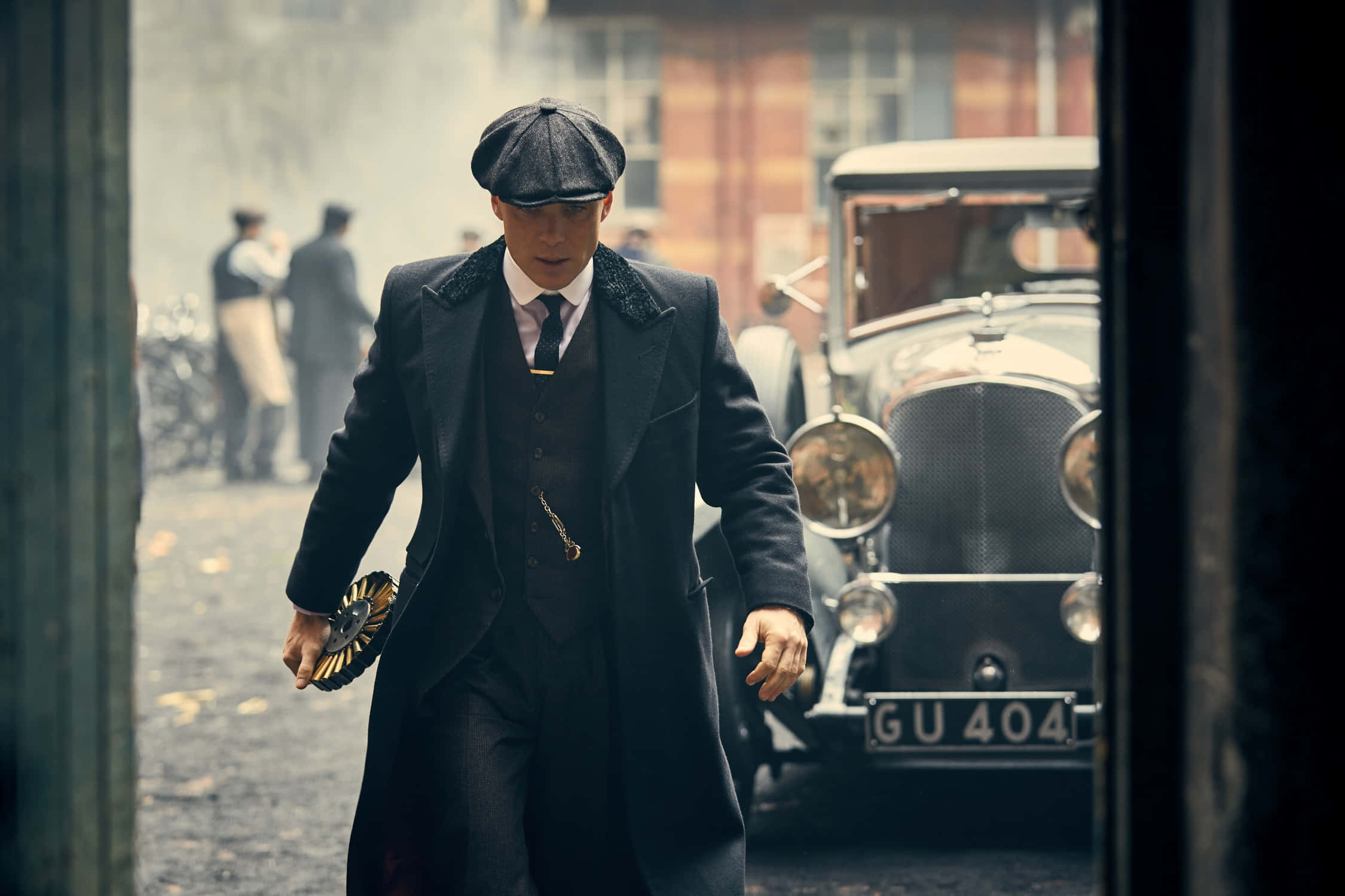 Peaky Blinders 桌布 壁紙 — 一名身穿深色西裝和扁帽的男子，在霧氣瀰漫的街道上，手持帽子，在一輛復古車旁大步向前。