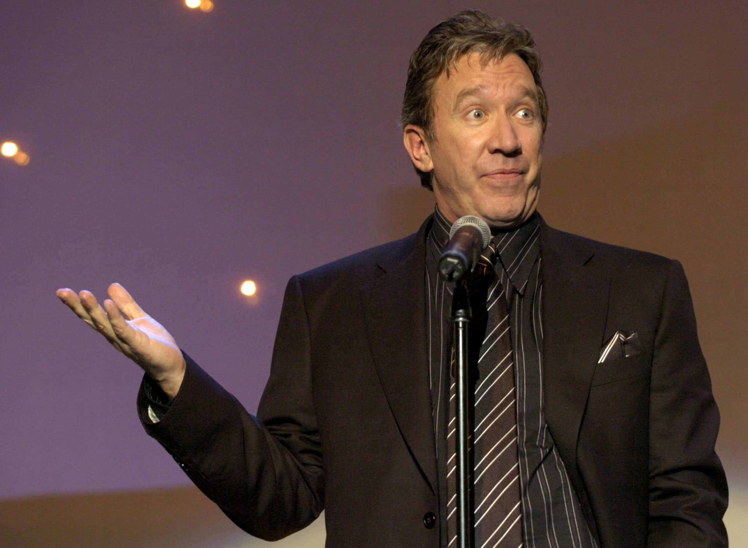 Tim Allen 桌布 壁紙 — 一名身穿深色西裝和條紋領帶的男子站在麥克風前，在舞台燈光下表情豐富，張開手勢。