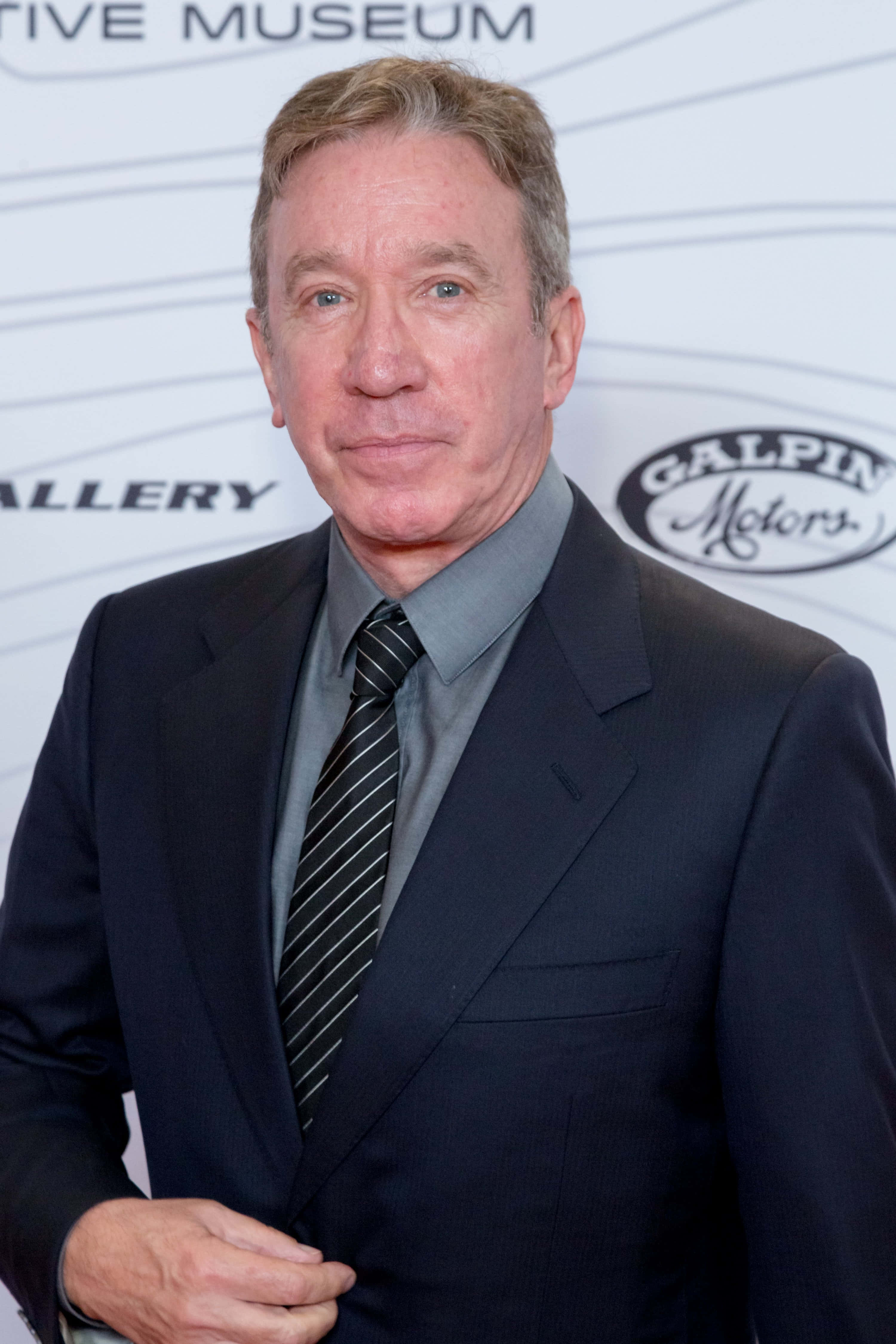 Tim Allen 桌布 壁紙 — 一名身穿深色西裝和條紋領帶的男子自信地站在博物館活動中，身後有標誌。