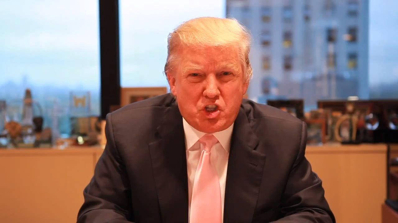 donald trump 桌布 壁紙 — 一名身穿深色西裝和粉紅色領帶的男子在室內坐下，背後是模糊的城市景觀，做出強烈的面部表情。