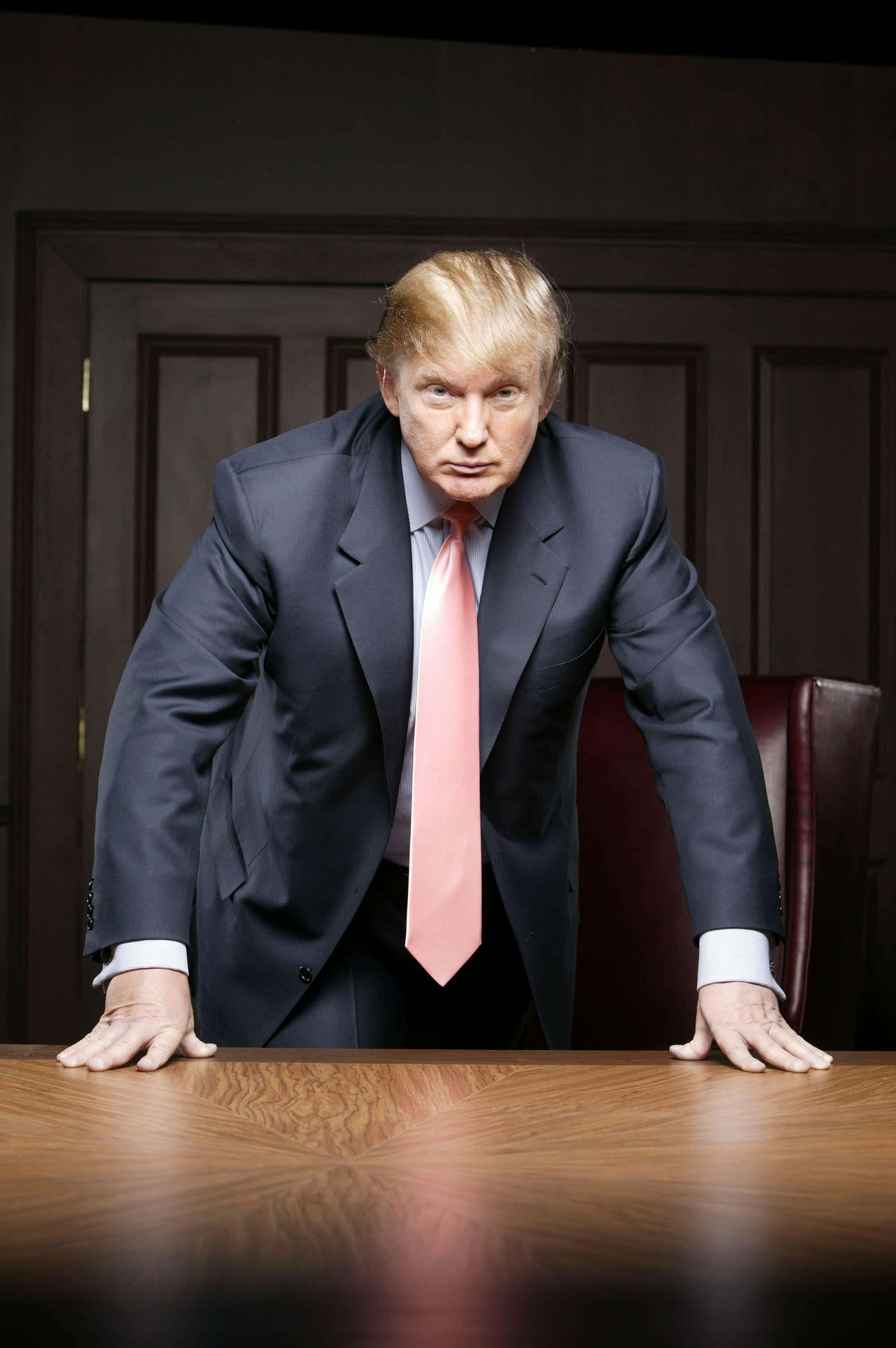 trump 桌布 壁紙 — 一名身穿深色西裝和粉紅色領帶的男子在燈光昏暗的辦公室裡，帶著強烈的表情俯身在一張木桌上。