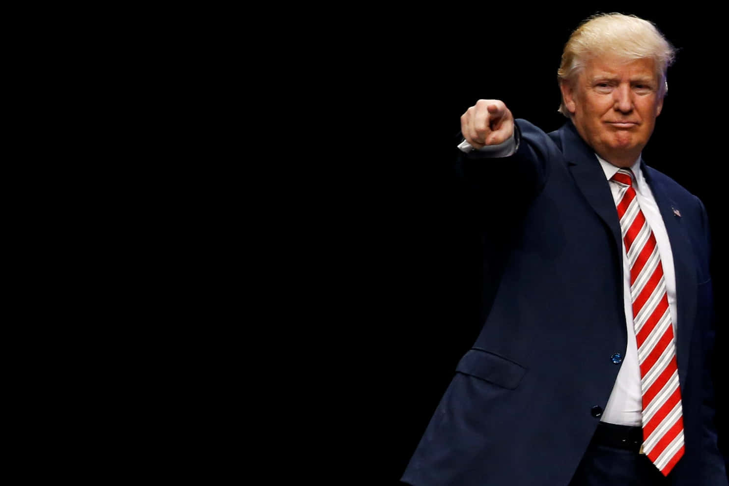 trump 桌布 壁紙 — 一名身穿深色西裝和紅色條紋領帶的男子在純黑背景前向前指。