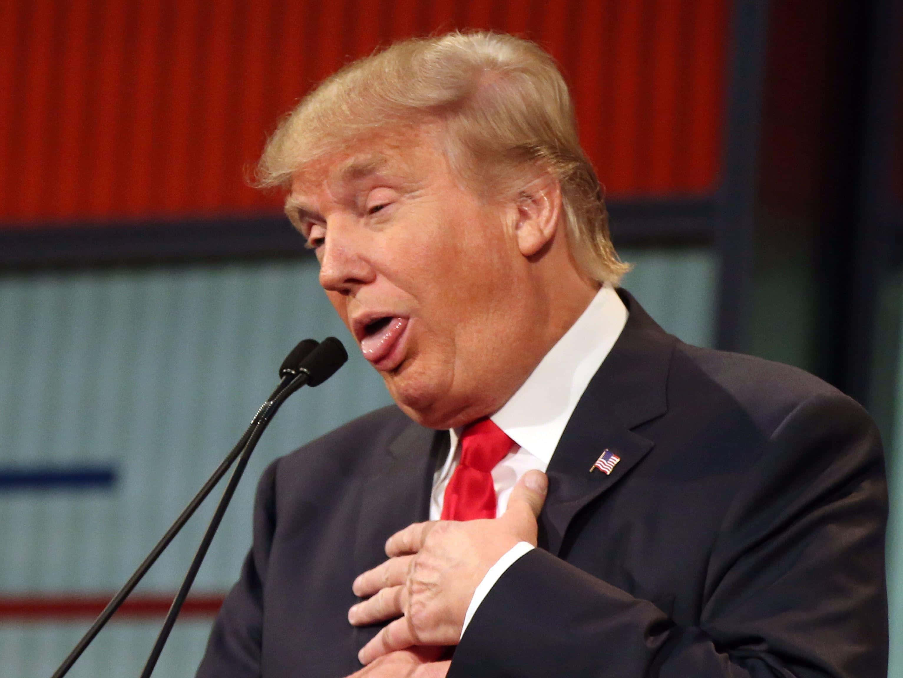 trump 桌布 壁紙 — 一名身穿深色西裝和紅色領帶的男子，在紅藍背景前，將手放在心口，熱情地對著麥克風說話。