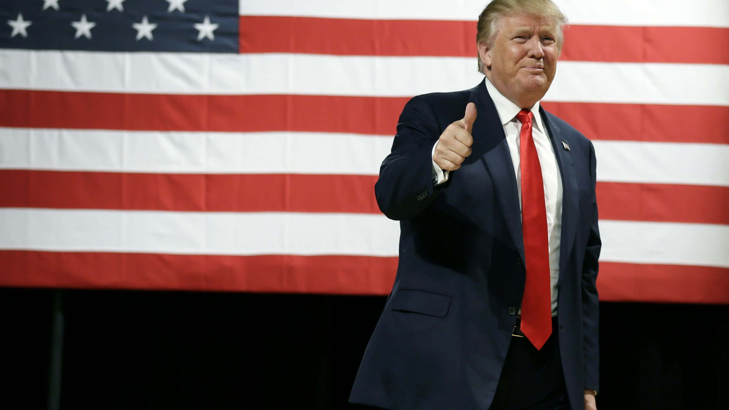 trump 桌布 壁紙 — 一名身穿深色西裝和紅色領帶的男子在大型美國國旗前豎起大拇指。
