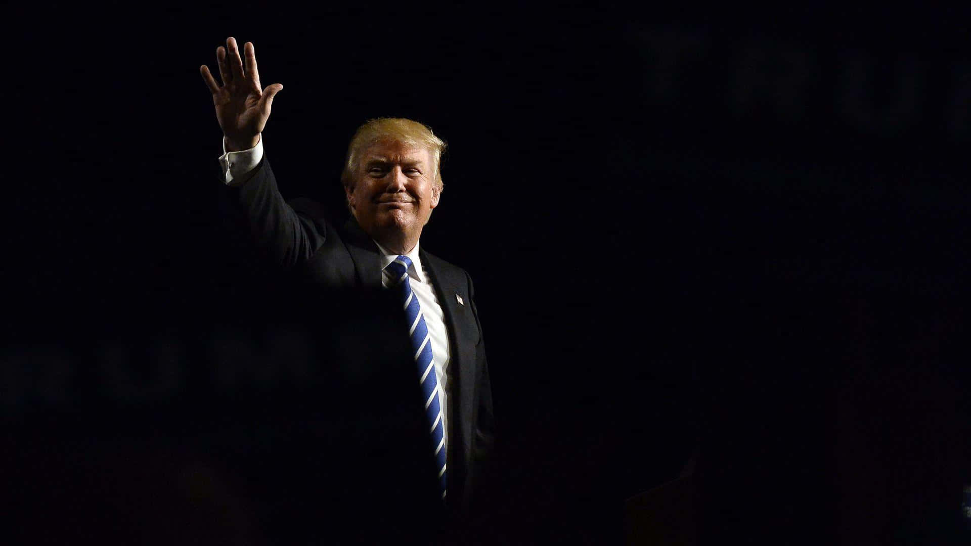 trump 桌布 壁紙 — 一名身穿深色西裝和藍色條紋領帶的男子在黑色背景前微笑揮手。