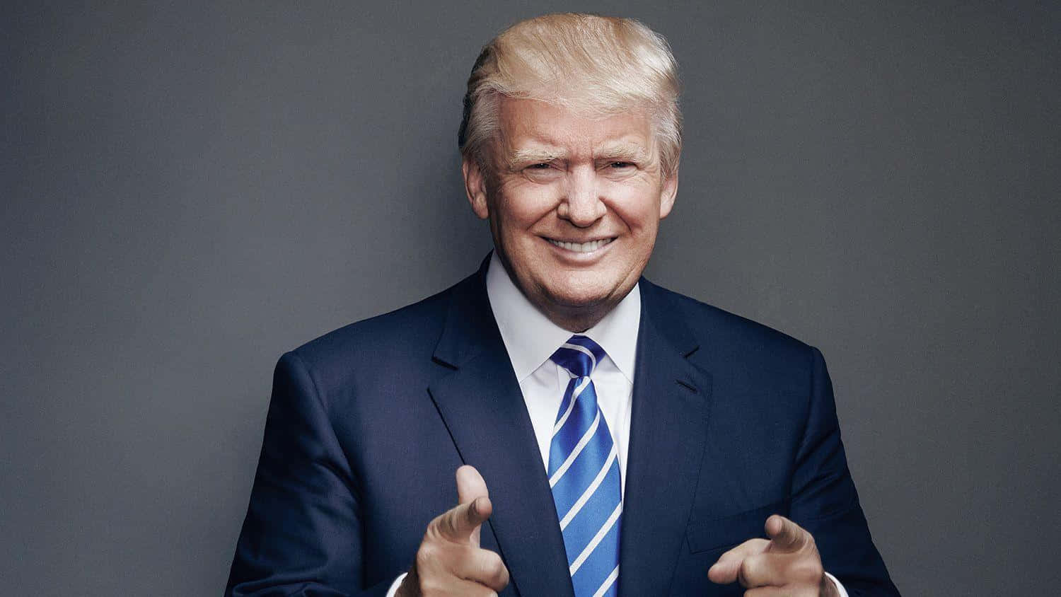 trump 桌布 壁紙 — 一名身穿深色西裝和藍色條紋領帶的男子，在純灰色背景前，雙手指向前方，露出燦爛的笑容。