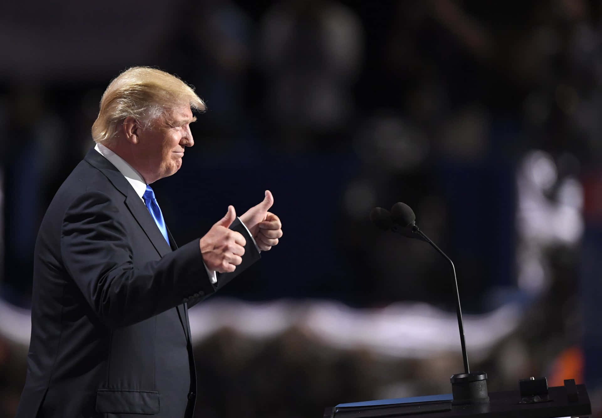 trump 桌布 壁紙 — 一名身穿深色西裝和藍色領帶的男子在燈光昏暗的舞台上站在講台前，背景是模糊的觀眾，他豎起大拇指。