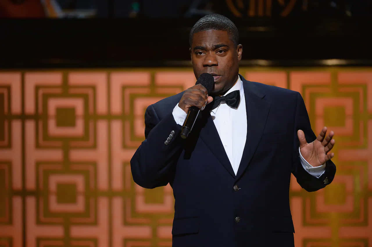 Tracy Morgan 桌布 壁紙 — 一名身穿深色西裝和領結的男子在幾何圖案背景的舞台上手持麥克風。